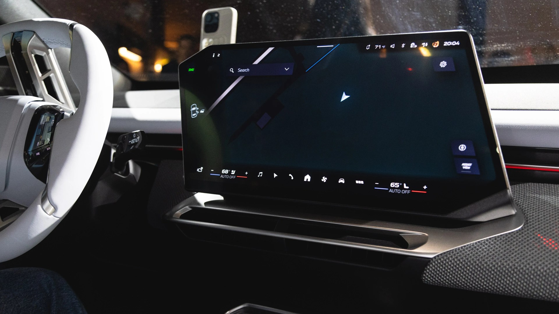 A modern car interior featuring a large, horizontally oriented touchscreen display on the dashboard. The screen shows a navigation interface with temperature controls at the bottom. The steering wheel is white with integrated buttons, and the dashboard has a sleek, minimalist design with a mix of light and dark materials.