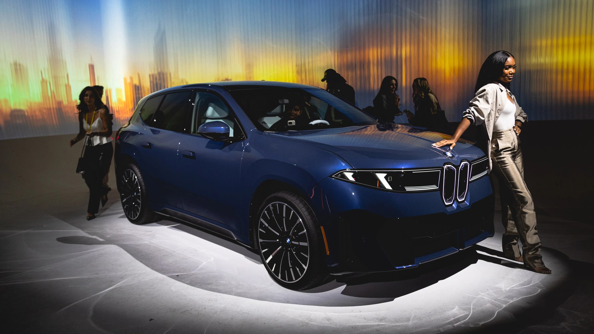 A dark blue BMW SUV displayed indoors with a spotlight on it, featuring sleek black alloy wheels and modern headlights. A woman dressed in a white top, beige jacket, and shiny beige pants is leaning against the front right side of the car. The background shows a blurred cityscape with warm sunset colors and several people in shadow.