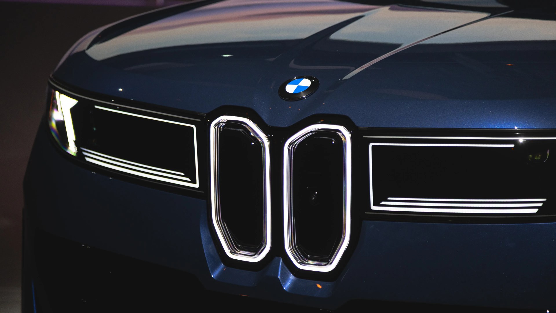 Front view of a dark blue BMW car featuring illuminated kidney grilles and sleek LED headlights with a modern, angular design. The BMW logo is centered above the grilles on the hood.