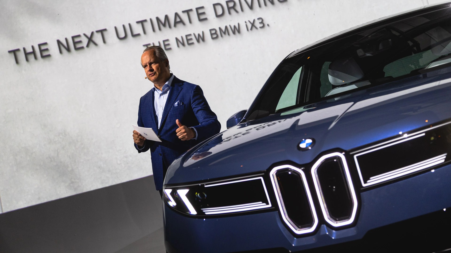 A man in a blue suit holding a card and giving a thumbs-up stands next to a blue BMW iX3 car. The background features a large screen with the text "THE NEXT ULTIMATE DRIVING" and "THE NEW BMW iX3." The car's front grille and headlights are prominently visible.