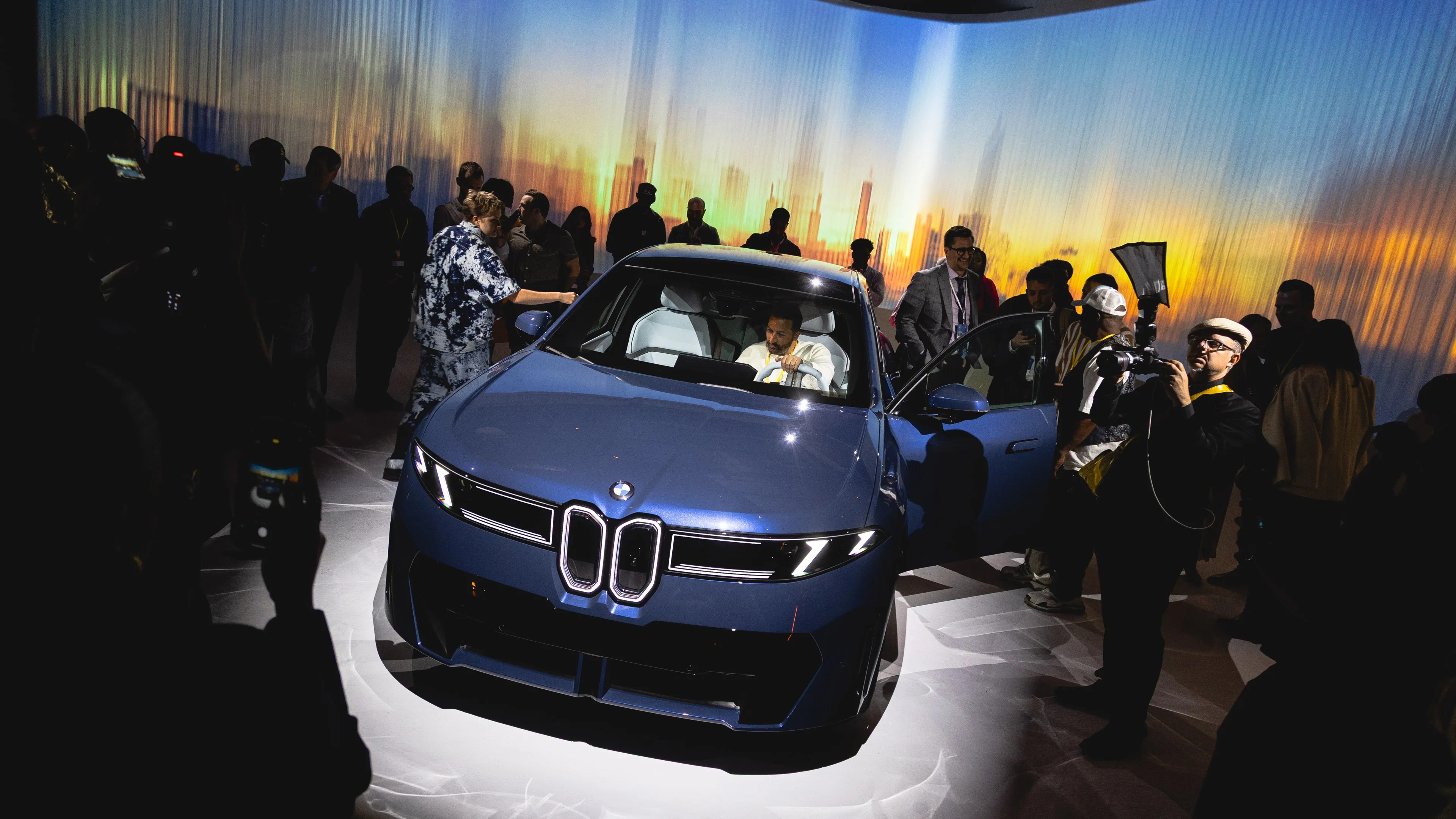 A blue BMW car is showcased in a dimly lit room with a crowd of people around it. The car's front is prominently visible, featuring sleek headlights and the iconic kidney grille. A man is seated inside the car, holding the steering wheel, while another person is reaching towards the driver's side mirror. The background displays a blurred cityscape with warm sunset colors. Several people are taking photos and observing the car.