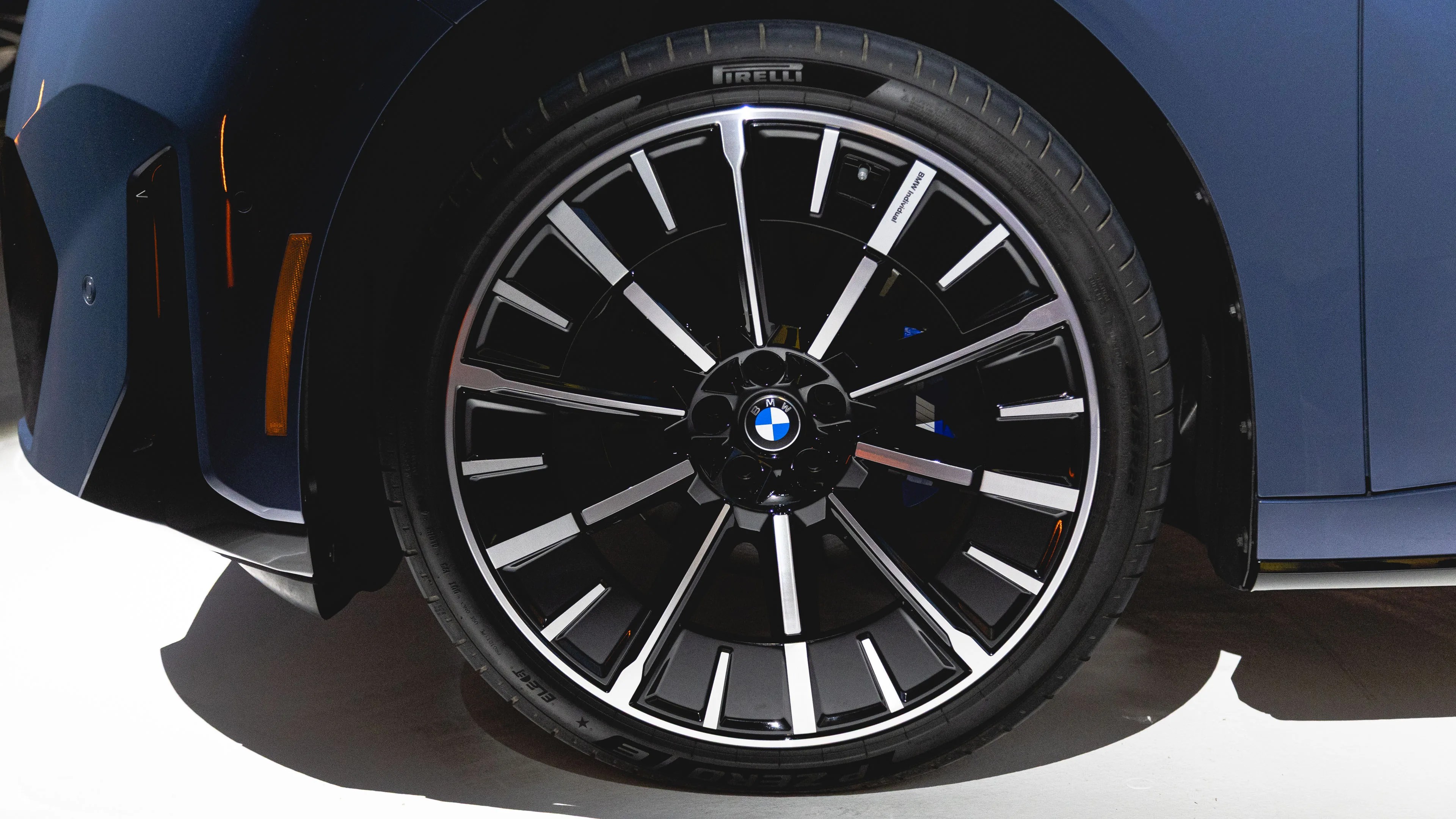 Close-up of a BMW car wheel featuring a black and silver multi-spoke alloy rim with the BMW logo in the center, fitted with a Pirelli P Zero tire. The car body is dark blue.