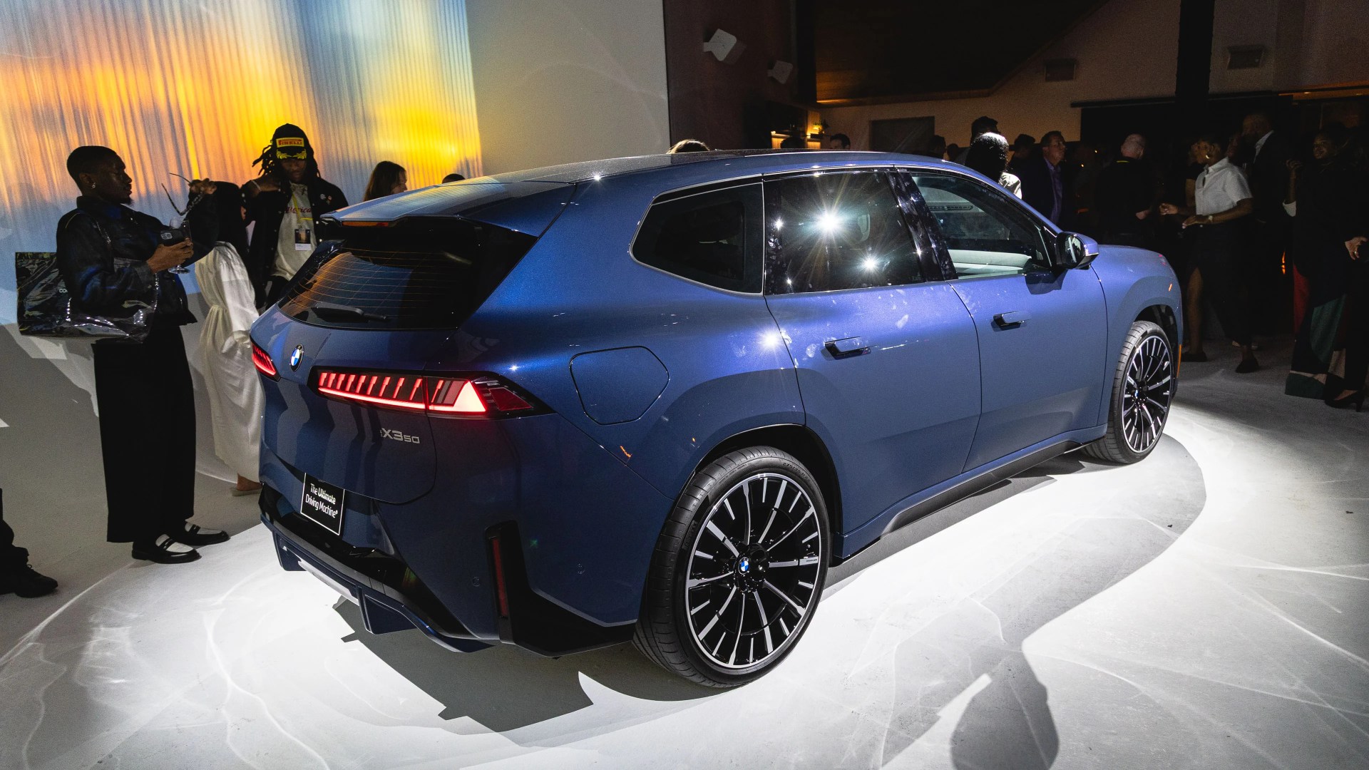 Blue BMW X3 50i SUV displayed indoors with people standing around, illuminated by bright spotlights highlighting its rear and side profile.