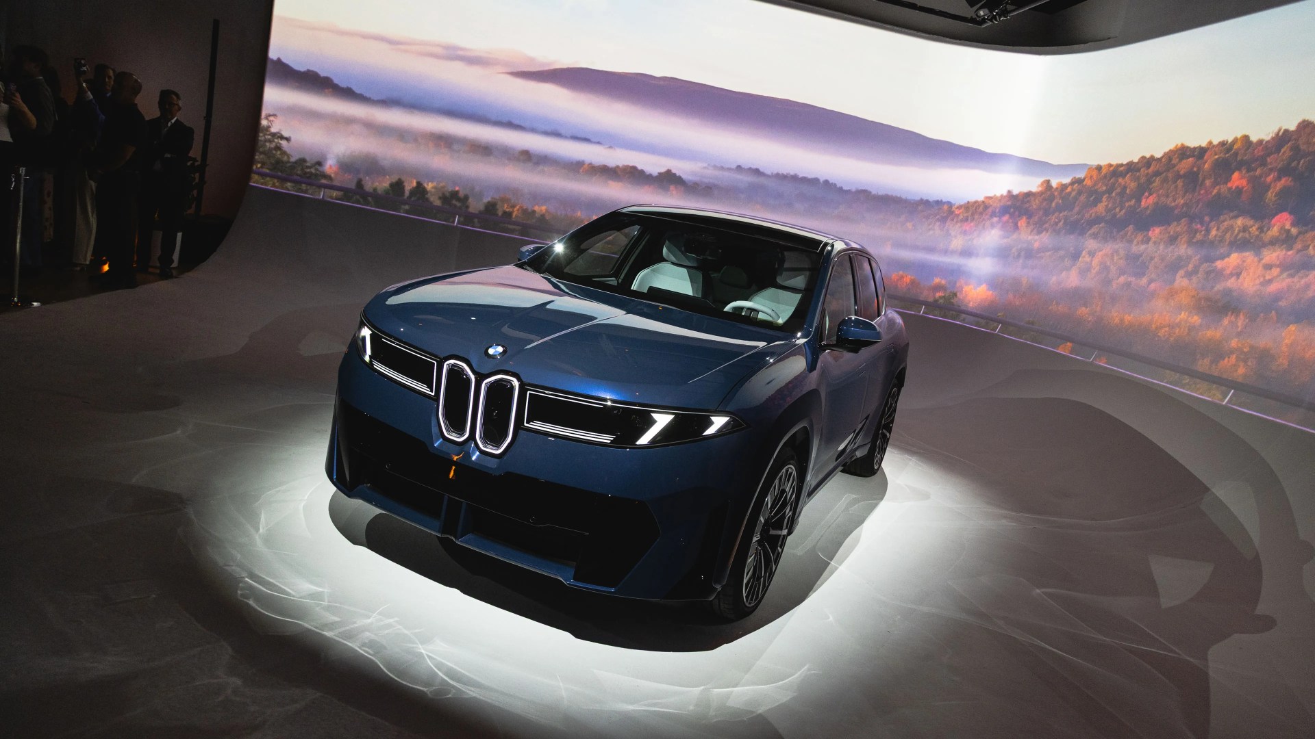 A dark blue BMW SUV is displayed under a spotlight in an indoor setting with a large scenic backdrop showing misty hills and autumn-colored trees. The car features a prominent kidney grille with illuminated outlines and sleek, modern headlights. The interior appears to have light-colored seats. Several people are visible in the dimly lit background on the left side.