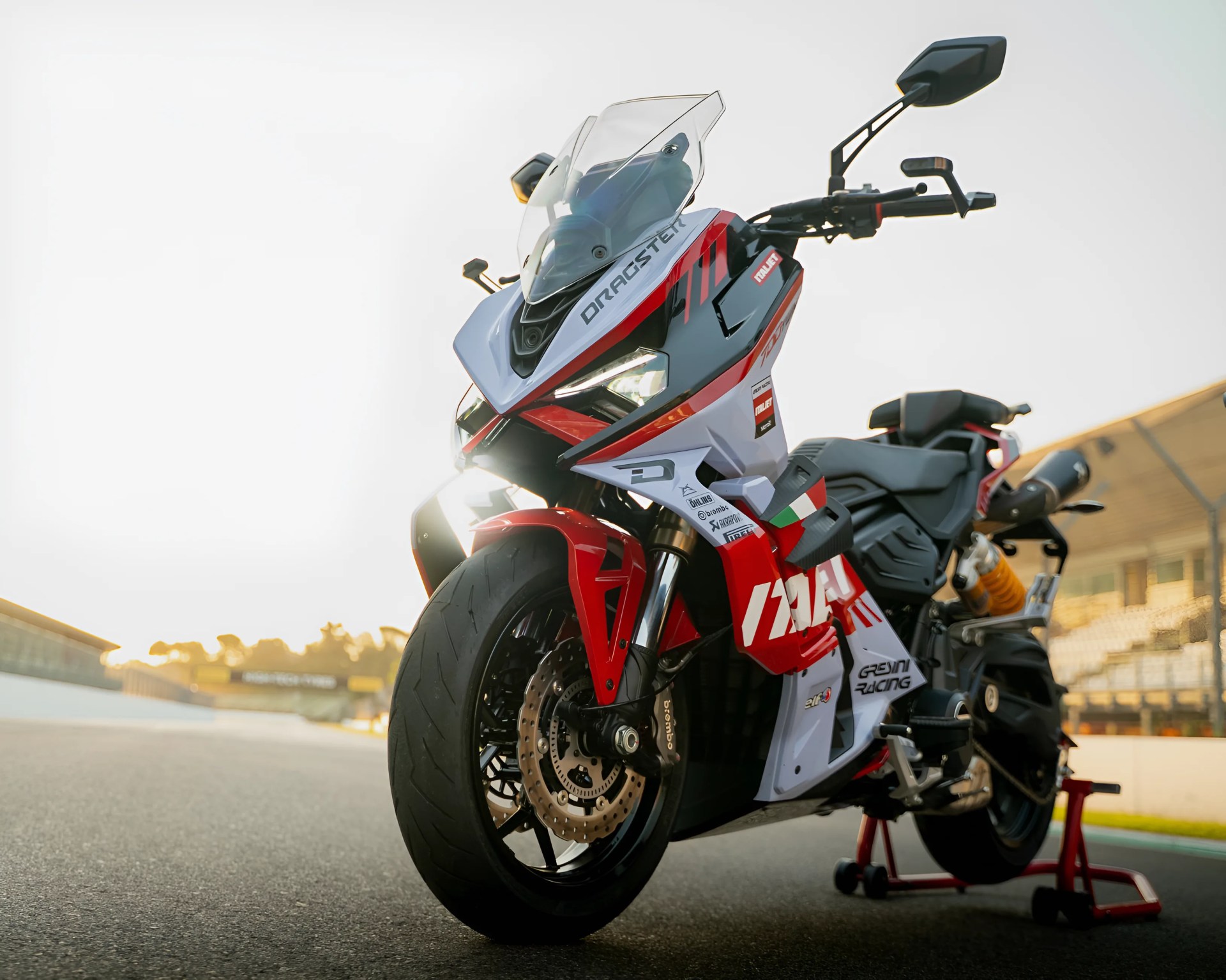 Sport motorcycle with a red, white, and black color scheme, featuring the word "DRAGSTER" on the front fairing. The bike has a clear windshield, dual front disc brakes, and is positioned on a rear stand on a racetrack with a blurred background of grandstands and fencing. The lighting suggests it is either early morning or late afternoon.