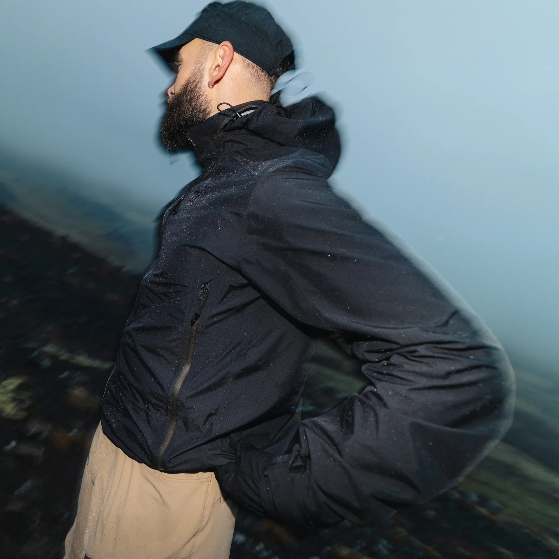 A man wearing a black cap, black zip-up jacket, and beige pants stands with his hands on his hips, looking to the left. The background is blurred, creating a motion effect. The man has a beard and an earring in his left ear.