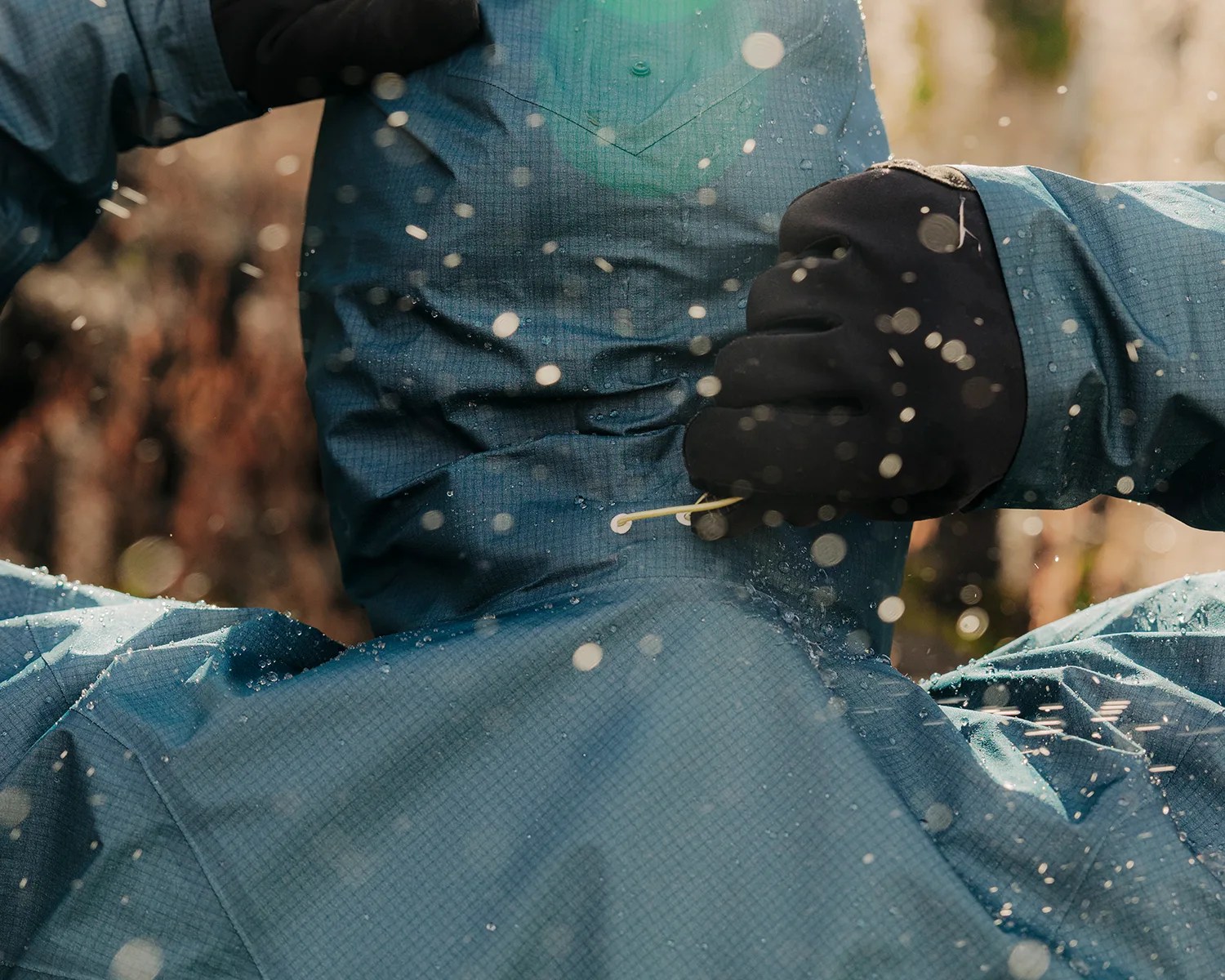 A person wearing a blue waterproof jacket and black gloves is pulling a drawstring on the jacket's hood. Water droplets are splashing around, indicating wet or rainy conditions. The background is blurred with natural outdoor colors.