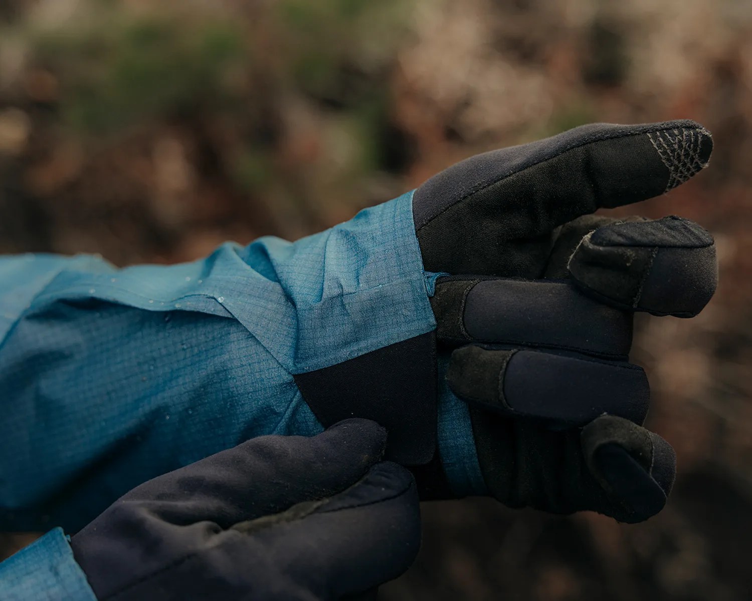 A close-up of a person wearing black gloves and a blue jacket, adjusting the cuff of the jacket. The gloves have reinforced stitching on the fingertips. The background is blurred with earthy tones.