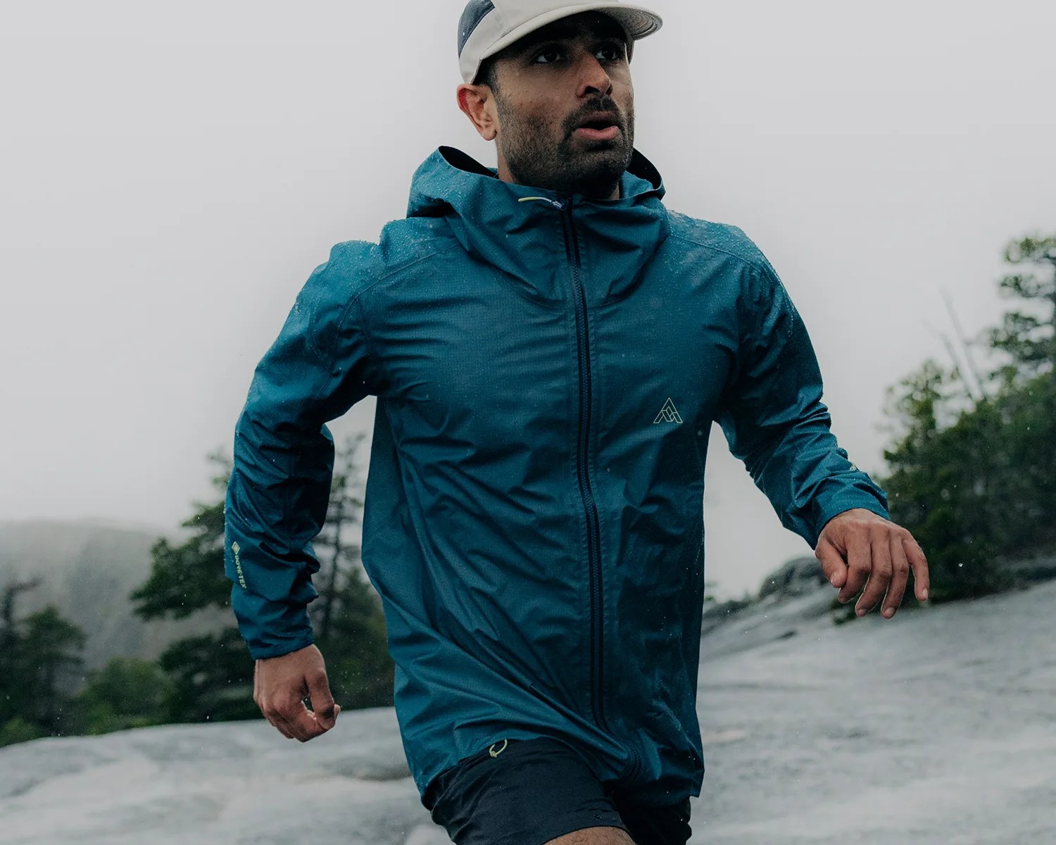 A man wearing a teal waterproof jacket with a hood and a light-colored cap, running outdoors on a wet, rocky surface with trees and a foggy sky in the background.
