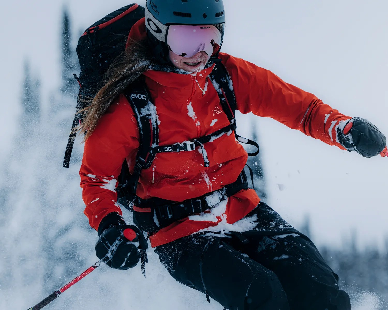 A person skiing in snowy conditions wearing a bright red jacket, black gloves, and black pants. They have a blue helmet with pink reflective goggles and a black backpack. Snow is visible on their clothing and in the air around them.
