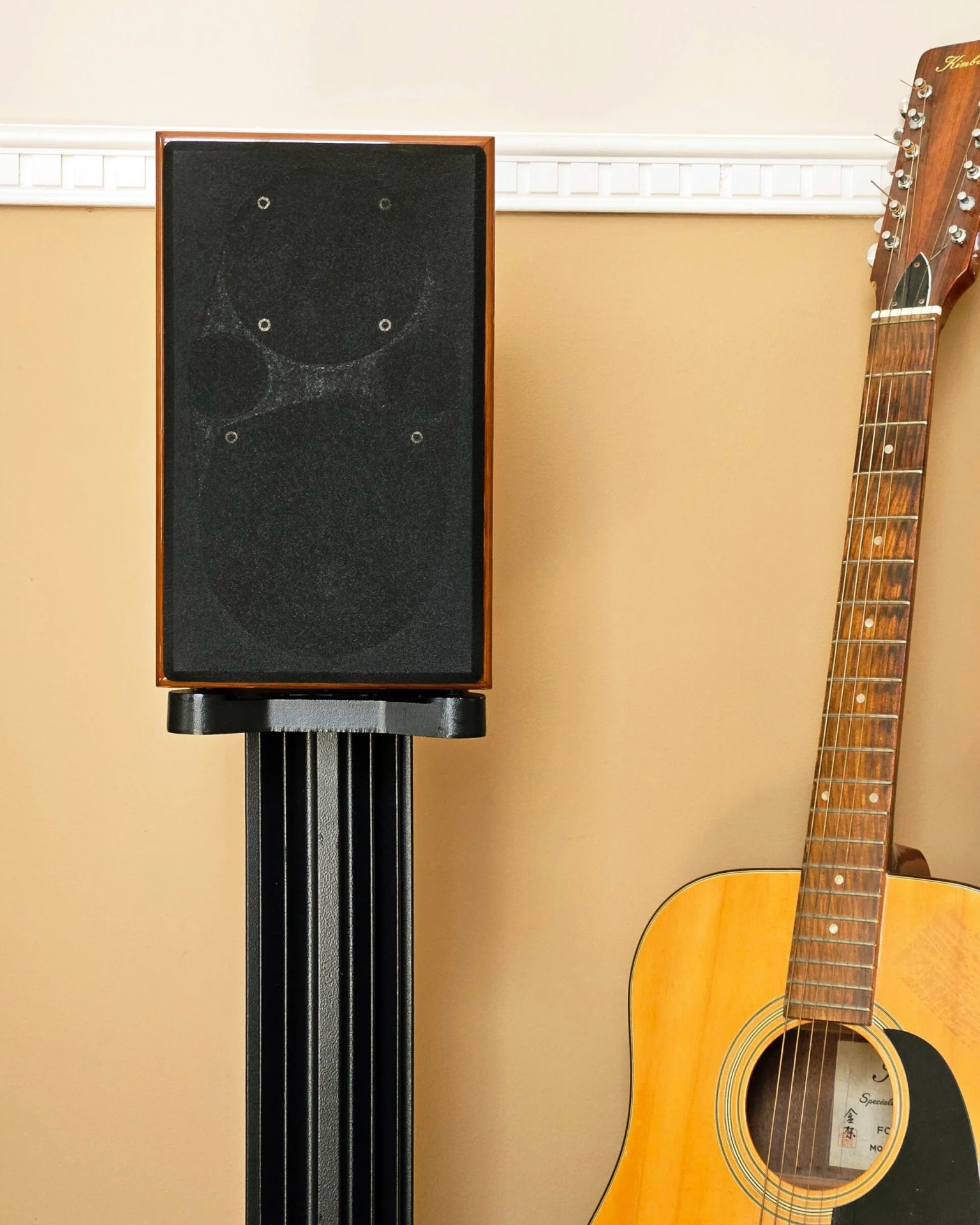 A wooden-framed speaker with a black fabric cover sits on a black stand next to an acoustic guitar with a natural wood finish and a dark pickguard. The background features a beige wall with white molding.