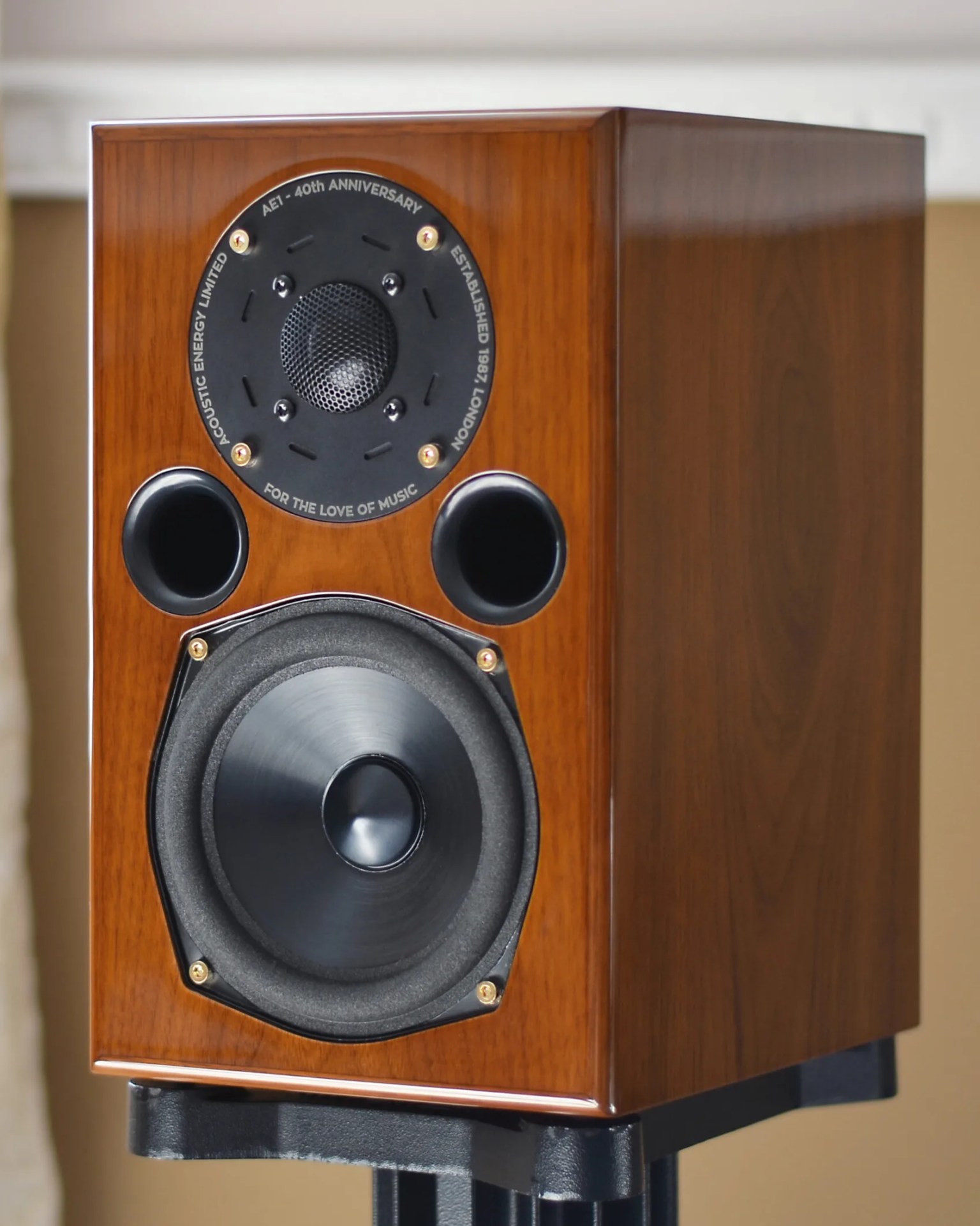 Wooden bookshelf speaker with a black tweeter, two circular ports, and a large black woofer mounted on a black stand. The tweeter has text around it that reads "AE1 - 40th ANNIVERSARY," "ACOUSTIC ENERGY LIMITED," "ESTABLISHED 1987, LONDON," and "FOR THE LOVE OF MUSIC.