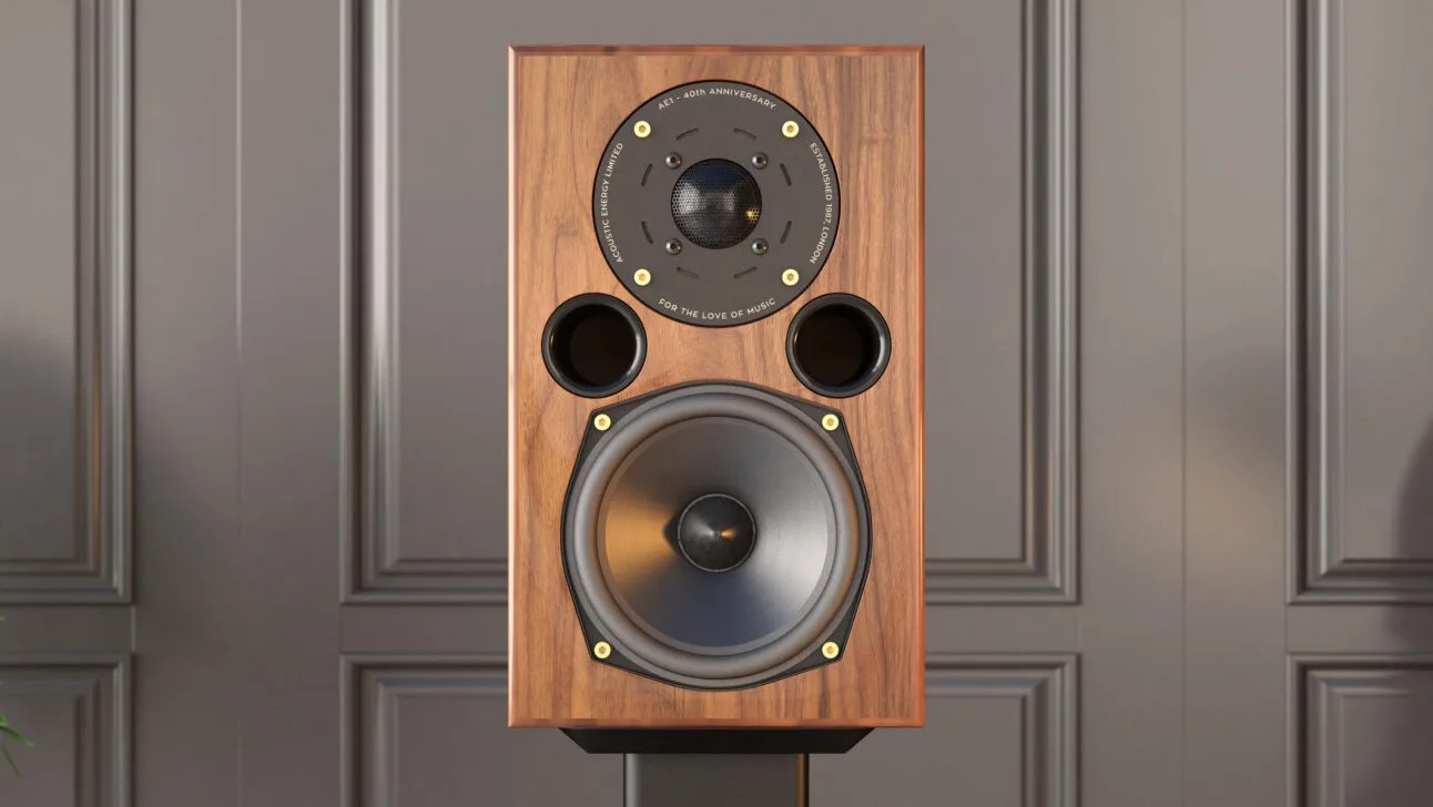 Wooden speaker with a large woofer at the bottom, two circular ports above it, and a tweeter at the top center. The tweeter is surrounded by a black circular plate with gold screws and white text that reads "AE1 - 40th Anniversary," "Acoustic Energy Limited," "Established 1987 London," and "For the Love of Music." The speaker is set against a gray paneled wall.