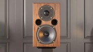 Wooden speaker with a large woofer at the bottom, two circular ports above it, and a tweeter at the top center. The tweeter is surrounded by a black circular plate with gold screws and white text that reads "AE1 - 40th Anniversary," "Acoustic Energy Limited," "Established 1987 London," and "For the Love of Music." The speaker is set against a gray paneled wall.