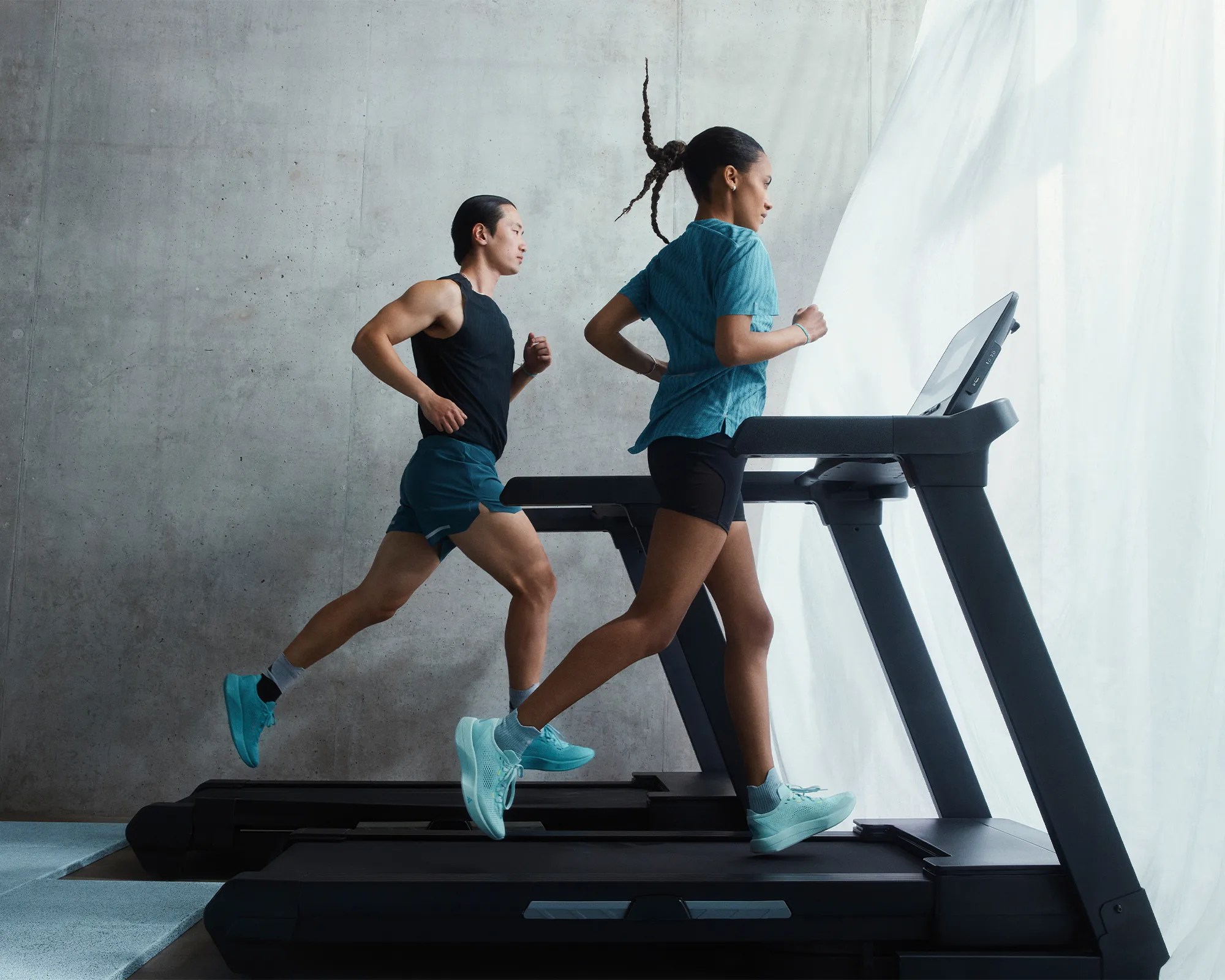 Two people running on treadmills in a modern indoor setting with a concrete wall and sheer white curtains in the background. The person in the foreground is wearing a blue shirt, black shorts, and light blue running shoes, while the person behind is wearing a black sleeveless top, dark shorts, and matching light blue running shoes.