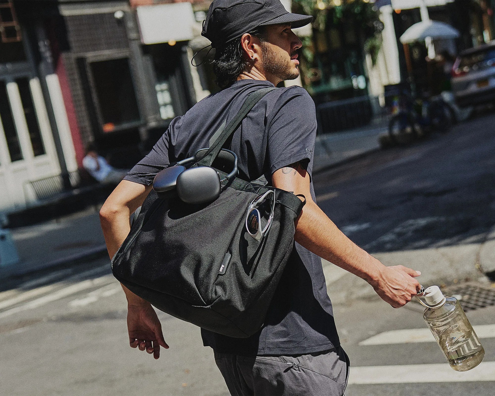 A man wearing a black cap and dark t-shirt is walking outdoors on a city street. He carries a large black shoulder bag with a pair of black headphones attached to it and sunglasses tucked into the side pocket. He holds a clear water bottle with a white cap in his right hand.