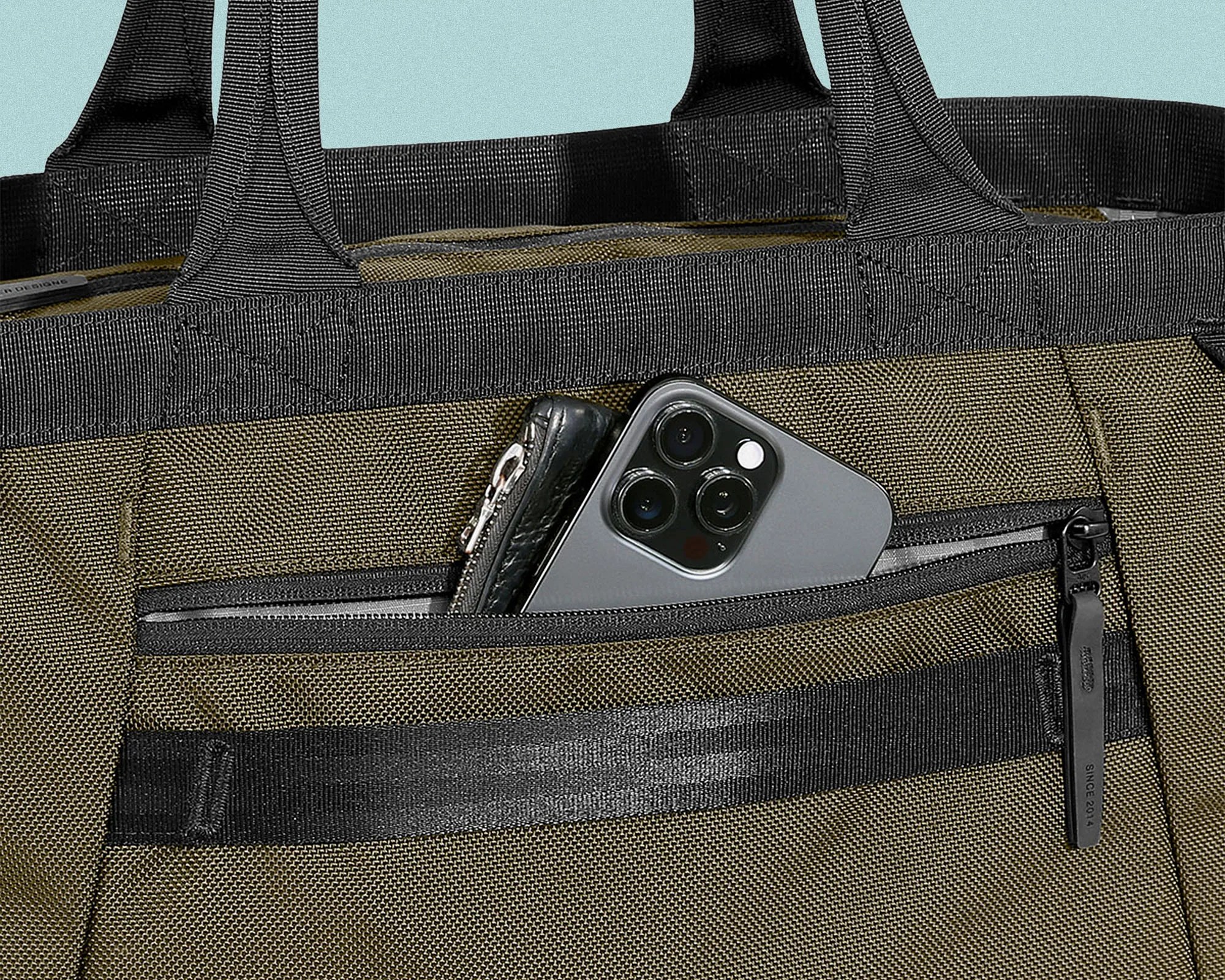 Close-up of an olive green and black fabric bag with a front zippered pocket partially open, containing a black leather wallet and a gray smartphone with three camera lenses. The bag has sturdy black handles and reinforced stitching.