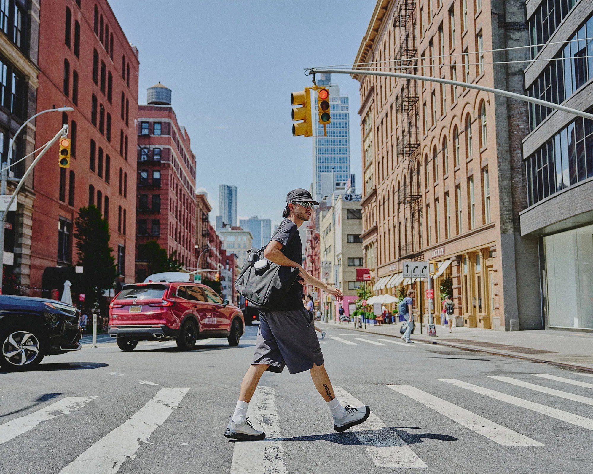 A man wearing a black cap, white sunglasses, a black t-shirt, and dark shorts is crossing a city street at a crosswalk. He carries a large black bag over his shoulder and wears white socks with white and black shoes. The street is lined with tall brick buildings, and there are cars waiting at a red traffic light. The sky is clear and blue.