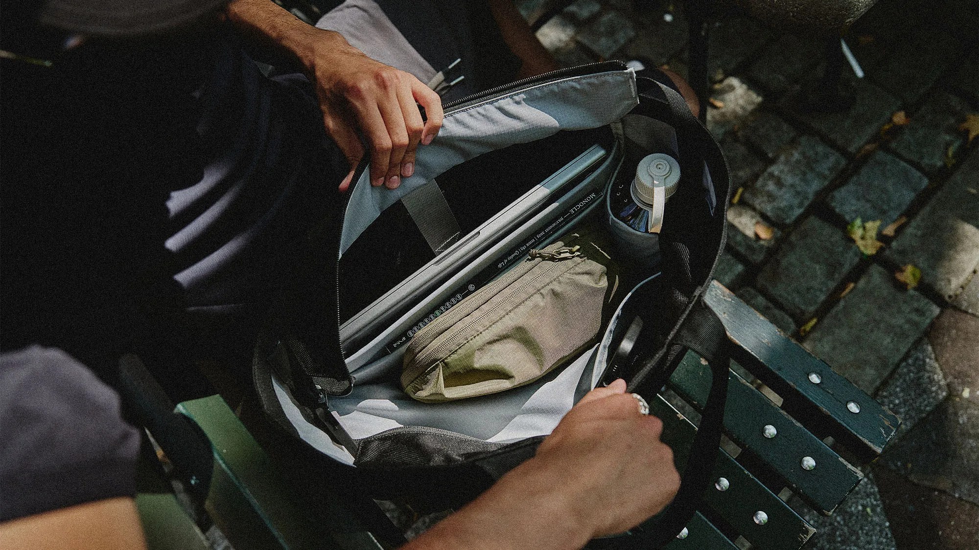 A black bag with a light gray interior is open, revealing a laptop, a beige zippered pouch, and a water bottle inside. The bag is resting on a green wooden bench with metal bolts, and a person's hands are holding the bag open. The background shows a stone-paved ground with scattered leaves.