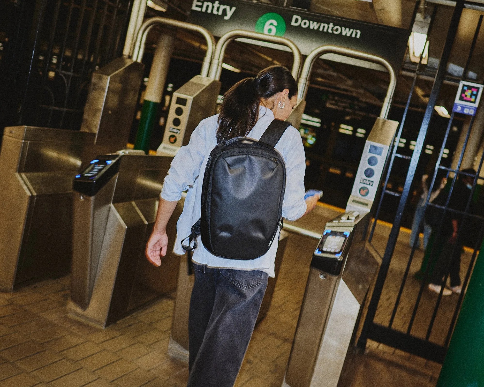 A person with long dark hair tied in a ponytail is wearing a light blue shirt, dark jeans, and a black backpack while using a smartphone to enter a subway station through a turnstile. The subway sign above reads "Entry 6 Downtown." The setting appears to be indoors with tiled flooring and metal turnstiles.