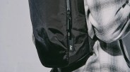 Close-up of a black backpack with a zipper pull tag labeled "Aer," worn over a white and gray plaid shirt. The fabric of the backpack has a slightly textured, durable appearance.