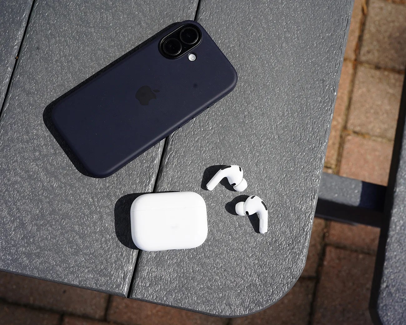 A black iPhone with a dual-camera setup, a white AirPods Pro charging case, and a pair of white AirPods Pro earbuds are placed on a textured gray outdoor table. The background includes a brick pavement.