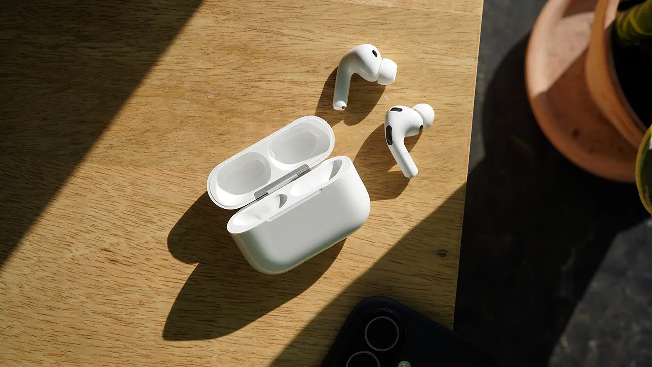 White wireless earbuds with an open charging case placed on a wooden surface, partially illuminated by sunlight.