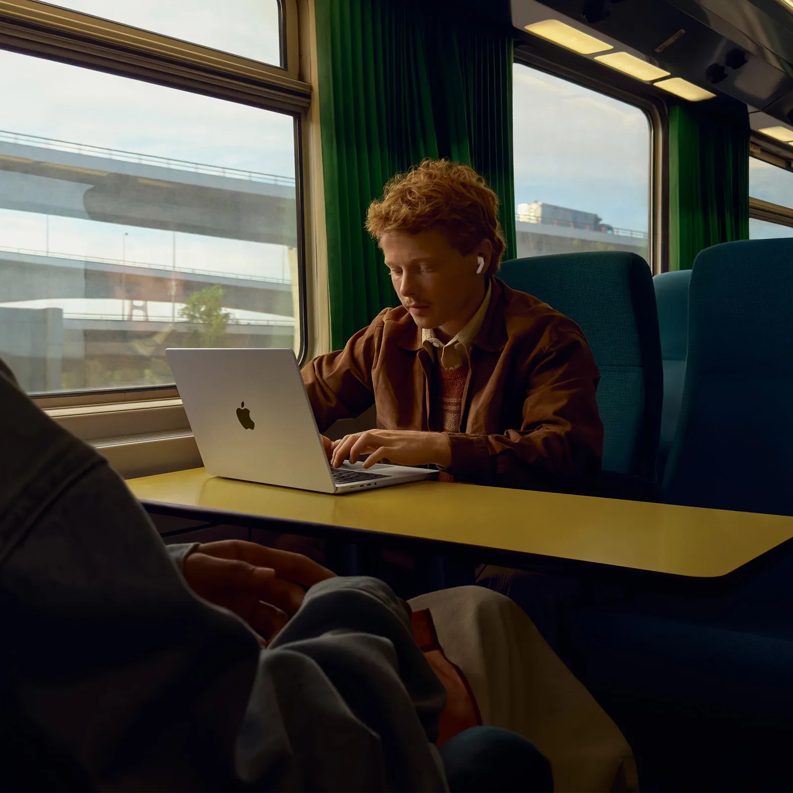 A person with curly red hair wearing a brown jacket and white earbuds is sitting at a yellow table inside a train, working on a silver Apple laptop. The train has large windows with green curtains and blue seats, and an overpass is visible outside. Another person, partially visible, is sitting nearby.