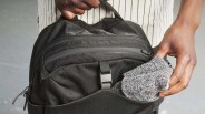 A close-up of a person placing a pair of gray knit socks into the front pocket of a black backpack. The person is wearing a white shirt with thin black vertical stripes. The backpack has multiple zippers and a black buckle on the front pocket.
