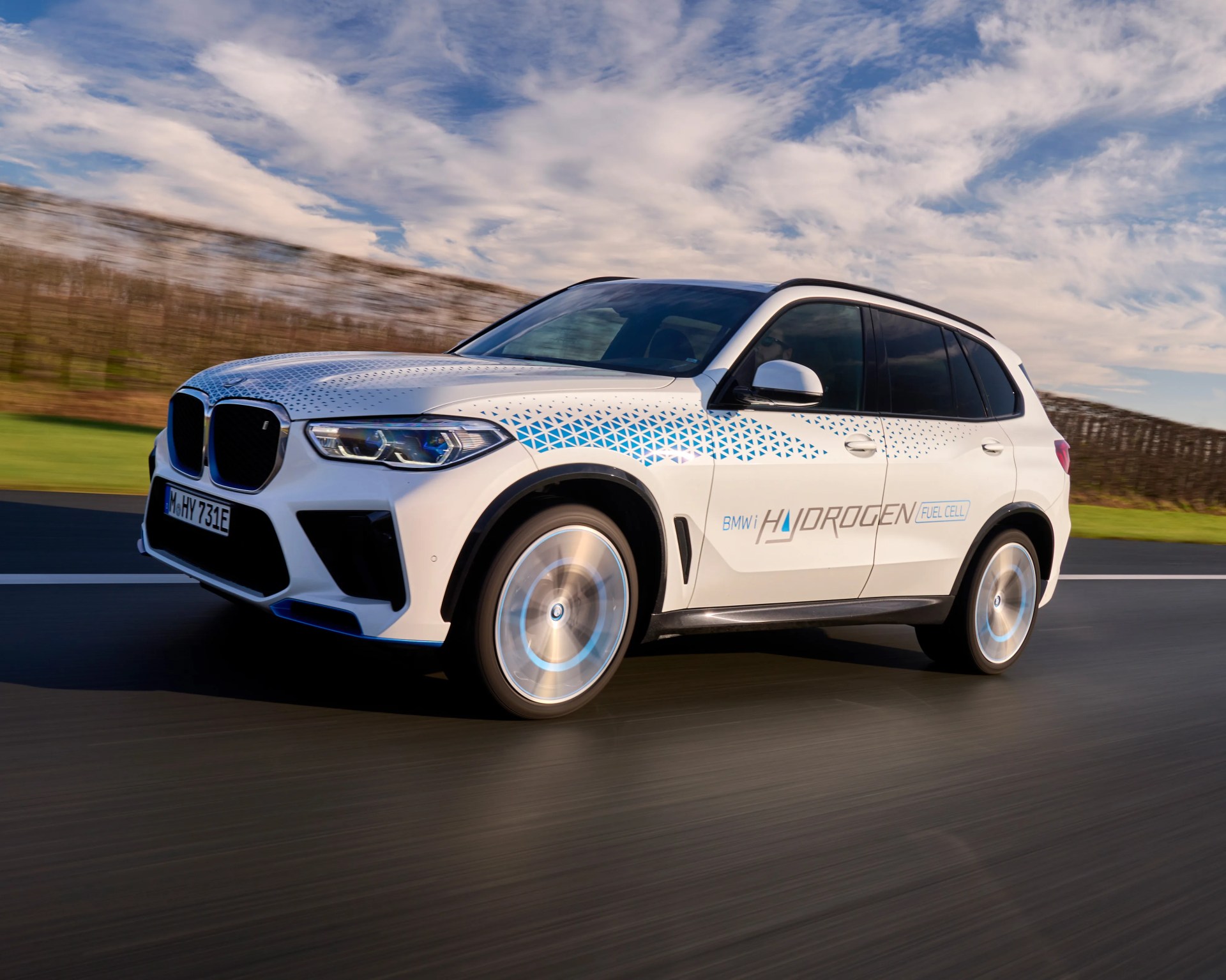 White BMW SUV with blue geometric patterns on the hood and sides, labeled "BMW i Hydrogen Fuel Cell," driving on a road with blurred background and partly cloudy sky.