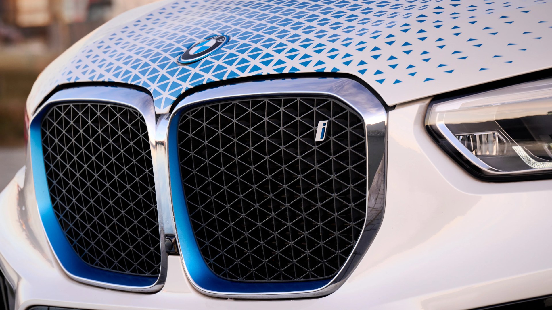 Close-up of the front grille of a white BMW car featuring the iconic kidney grille design with a blue gradient trim and a geometric pattern on the hood. The right grille has a small "i" badge, indicating it is a BMW i model. The right headlight is partially visible with a modern, angular design.