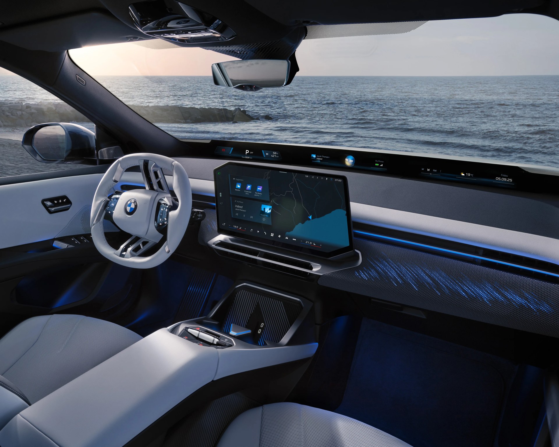 Interior of a modern BMW car featuring a white and black steering wheel with the BMW logo, a large central touchscreen display showing navigation and media controls, and a sleek dashboard with a wide digital instrument cluster extending across the front. The cabin has ambient blue lighting and light-colored seats, with a view of the ocean and rocky shore visible through the windows.