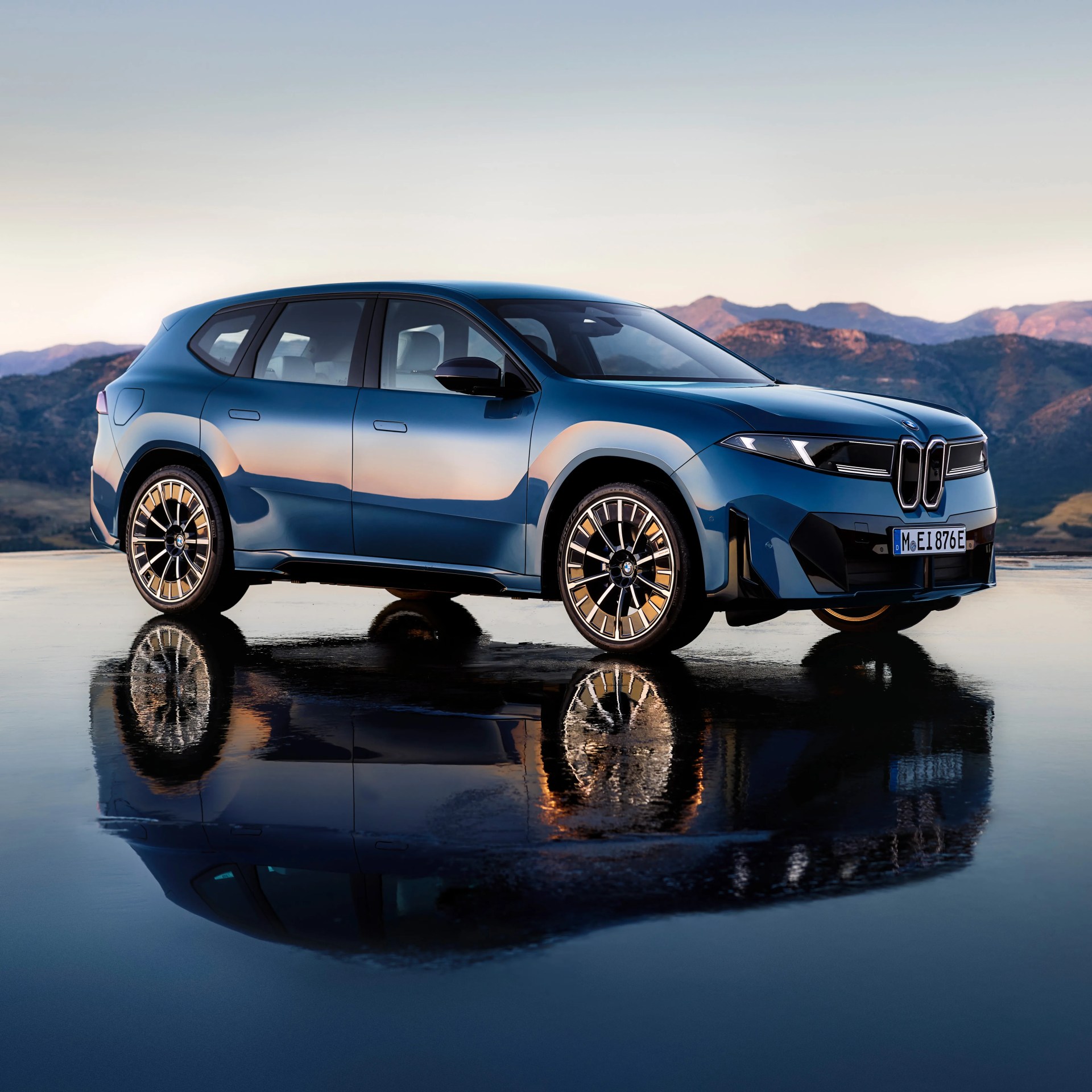 Blue BMW electric SUV parked on a reflective wet surface with mountainous landscape in the background during sunset.