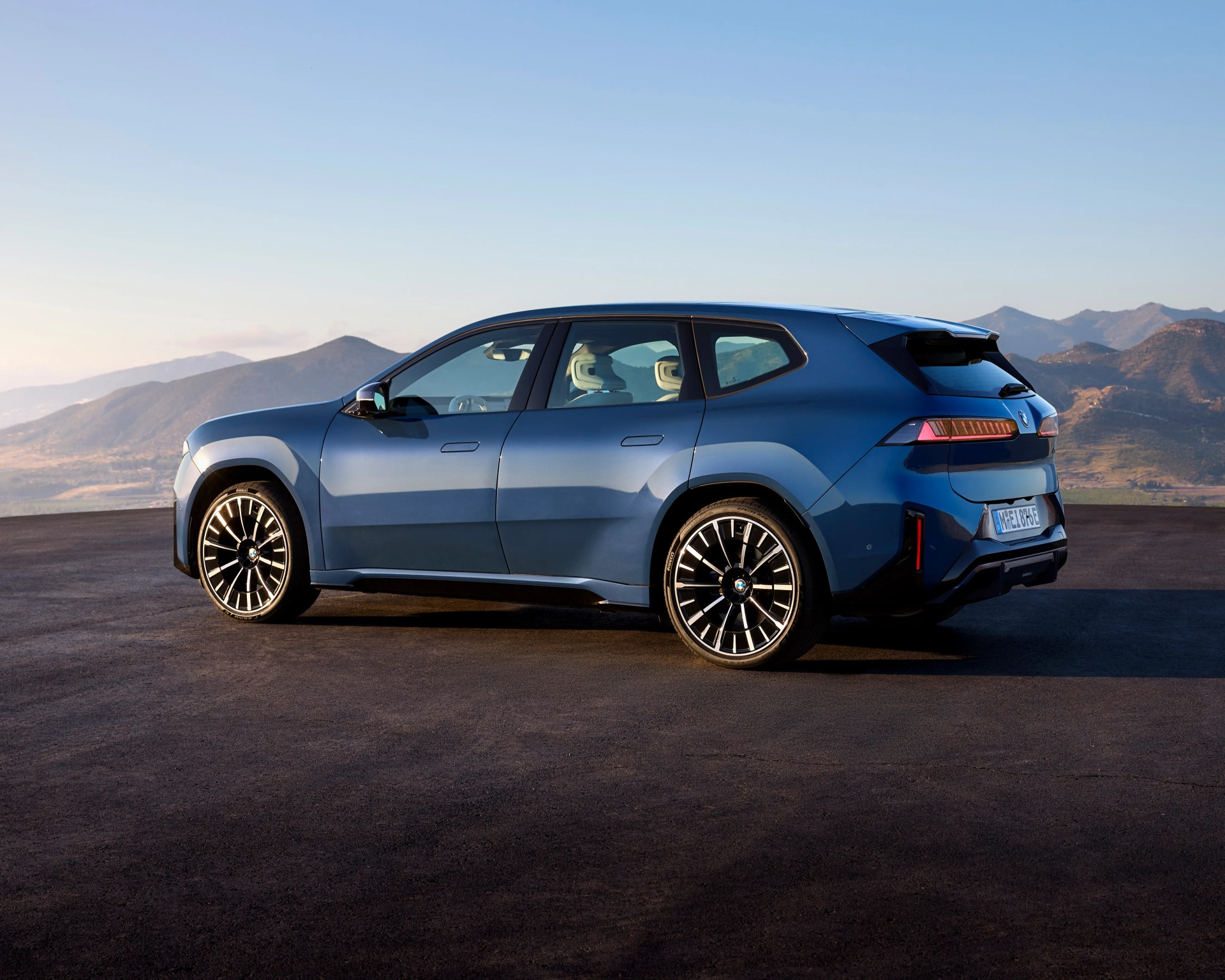 Blue BMW SUV with sleek, modern design, black and silver multi-spoke wheels, and tinted rear windows, parked on a flat asphalt surface with mountainous landscape in the background under a clear sky.