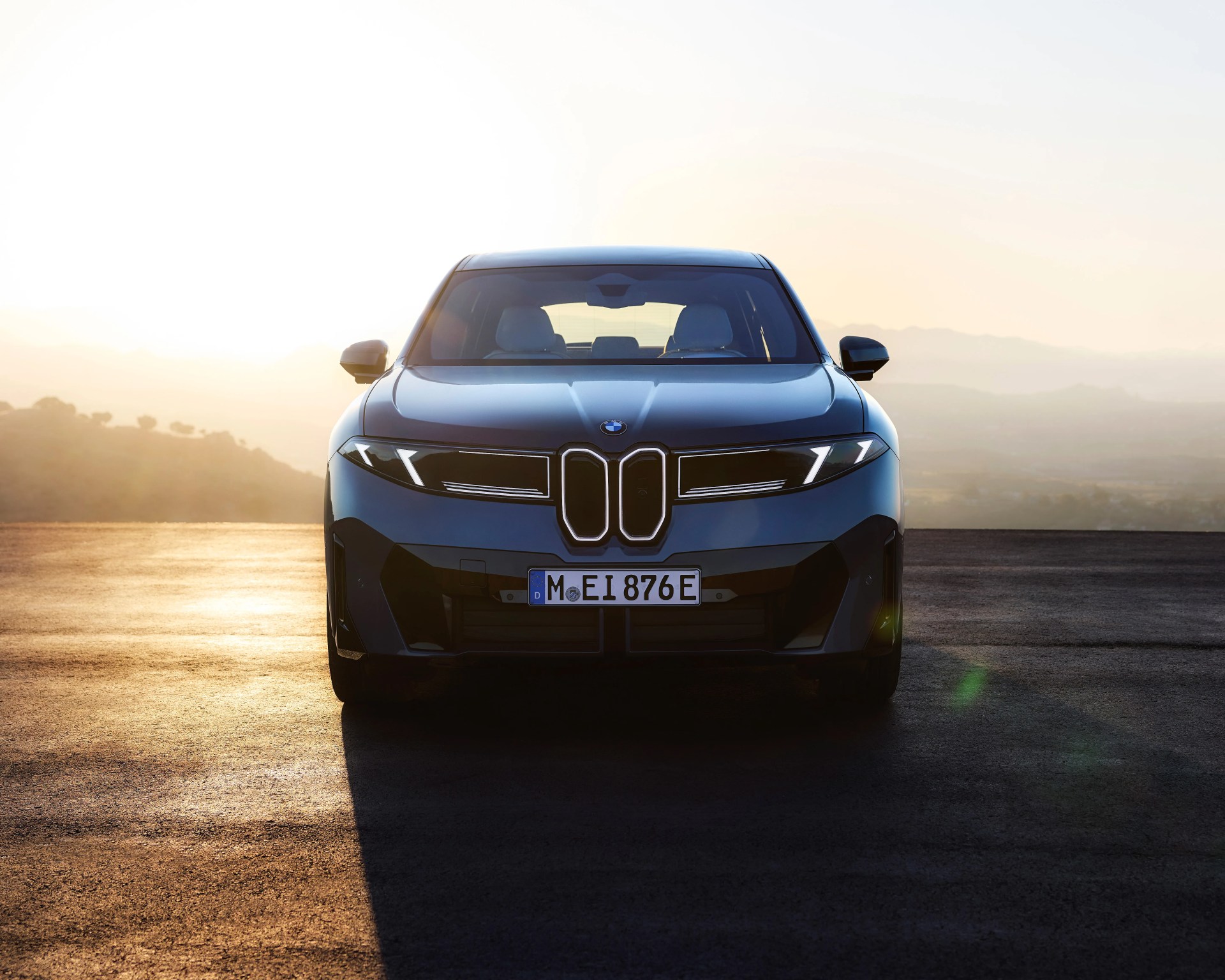 Front view of a sleek, modern BMW car with distinctive kidney-shaped grille and sharp LED headlights, parked on an asphalt surface with a hazy, mountainous landscape in the background during sunrise or sunset.