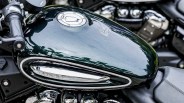 Glossy dark green motorcycle fuel tank with a silver fuel cap and a chrome accent piece on the side, reflecting trees and sky. The word "BENGAL" is faintly visible on the chrome accent. The surrounding parts of the motorcycle include black handlebars and a black seat.