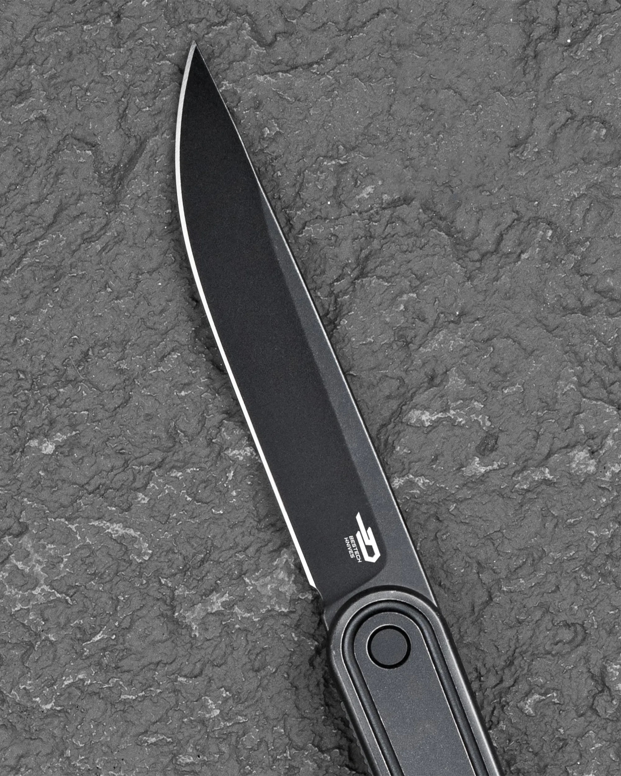 Black folding knife blade with a sleek, matte finish and a sharp edge, resting on a textured dark gray surface. The blade has a logo and text near the handle that reads "BESTECH KNIVES." The handle is partially visible, featuring a circular button or pivot point.