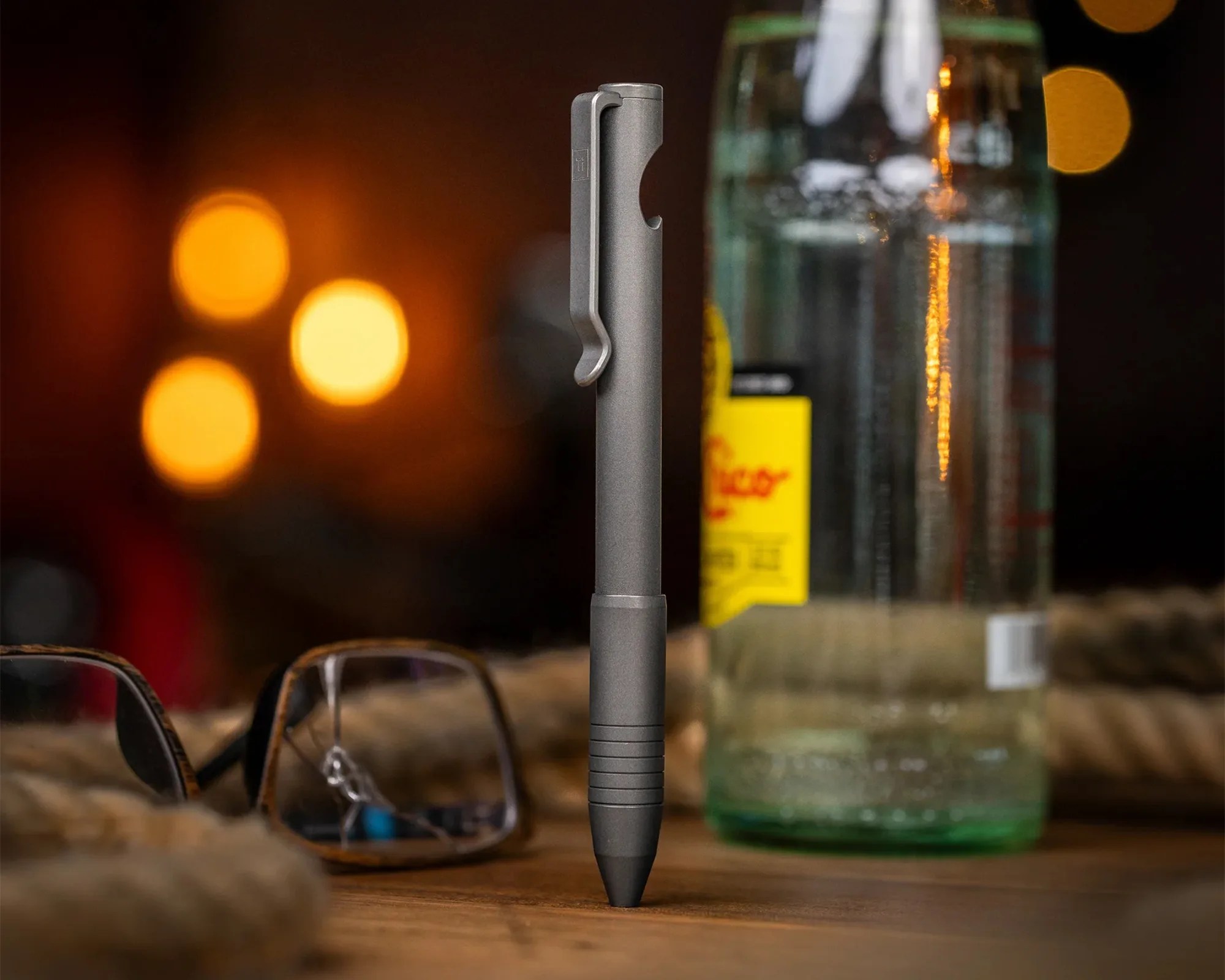 A sleek, metallic gray pen standing upright on a wooden surface. The pen has a clip near the top and a textured grip area near the tip. In the background, there is a pair of eyeglasses lying on the surface and a glass bottle with a yellow label, both slightly out of focus. Warm, circular bokeh lights add a cozy ambiance to the scene.