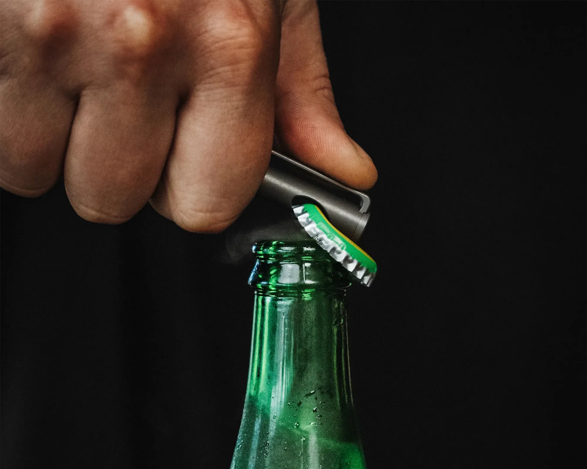 A hand is using a metal bottle opener to remove a green and yellow bottle cap from a green glass bottle against a black background.
