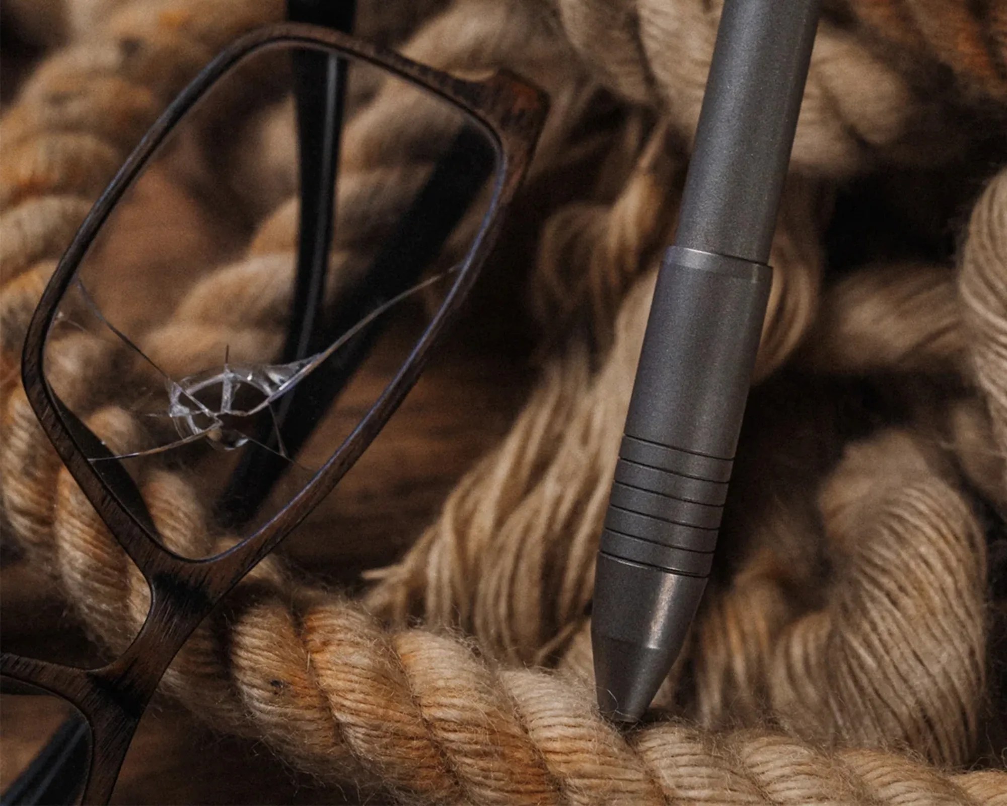 A close-up of a pair of eyeglasses with a cracked lens resting on thick, twisted beige rope. Next to the glasses is a dark gray pen with a textured grip.