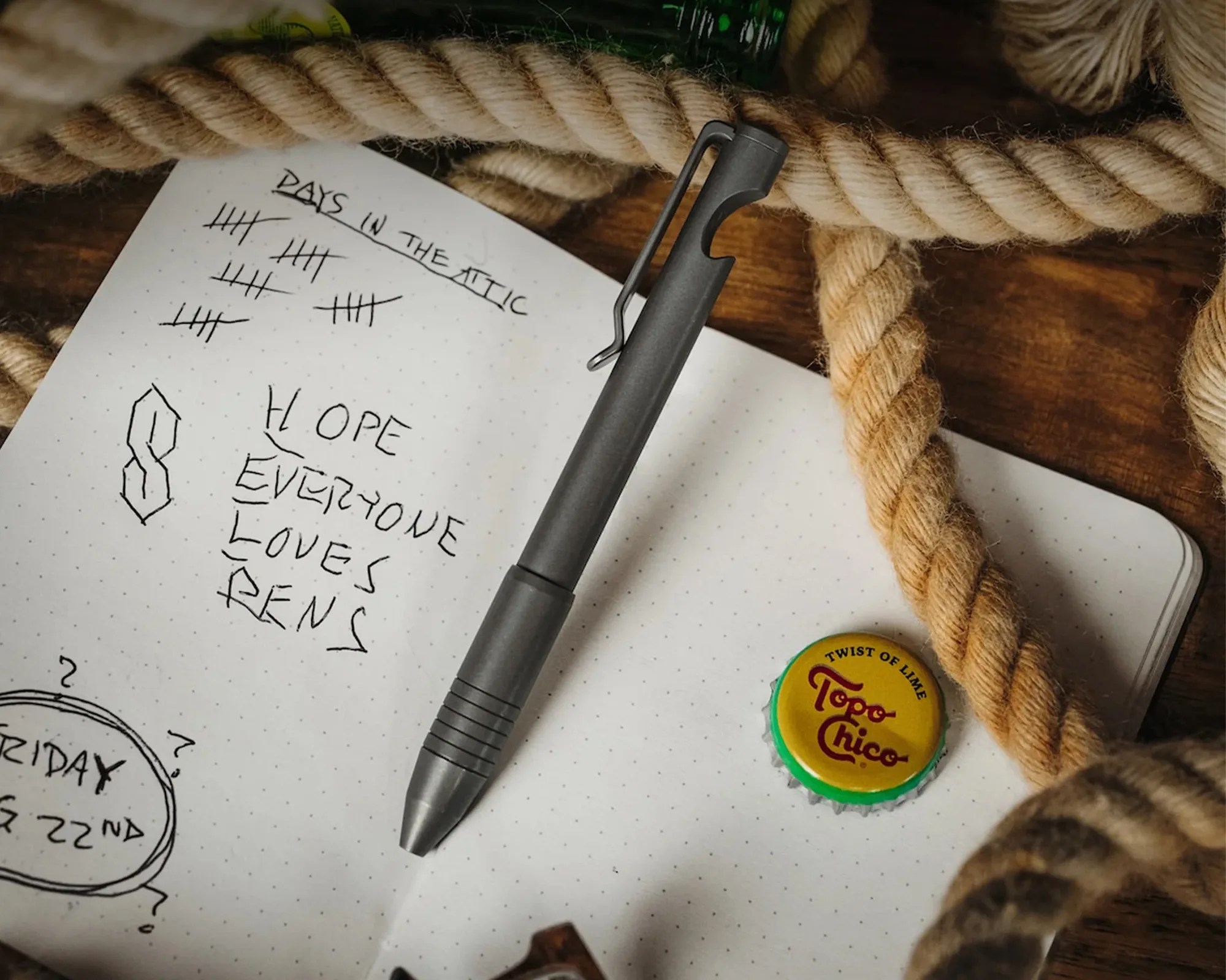 A gray pen rests on an open dotted notebook page with handwritten text and tally marks. The text reads "DAYS IN THE ATTIC" with tally marks underneath, and "HOPE EVERYONE LOVES PENS" written below a stylized "S" symbol. A yellow and green Topo Chico bottle cap labeled "TWIST OF LIME" is also on the notebook. Thick beige ropes surround the notebook on a wooden surface.