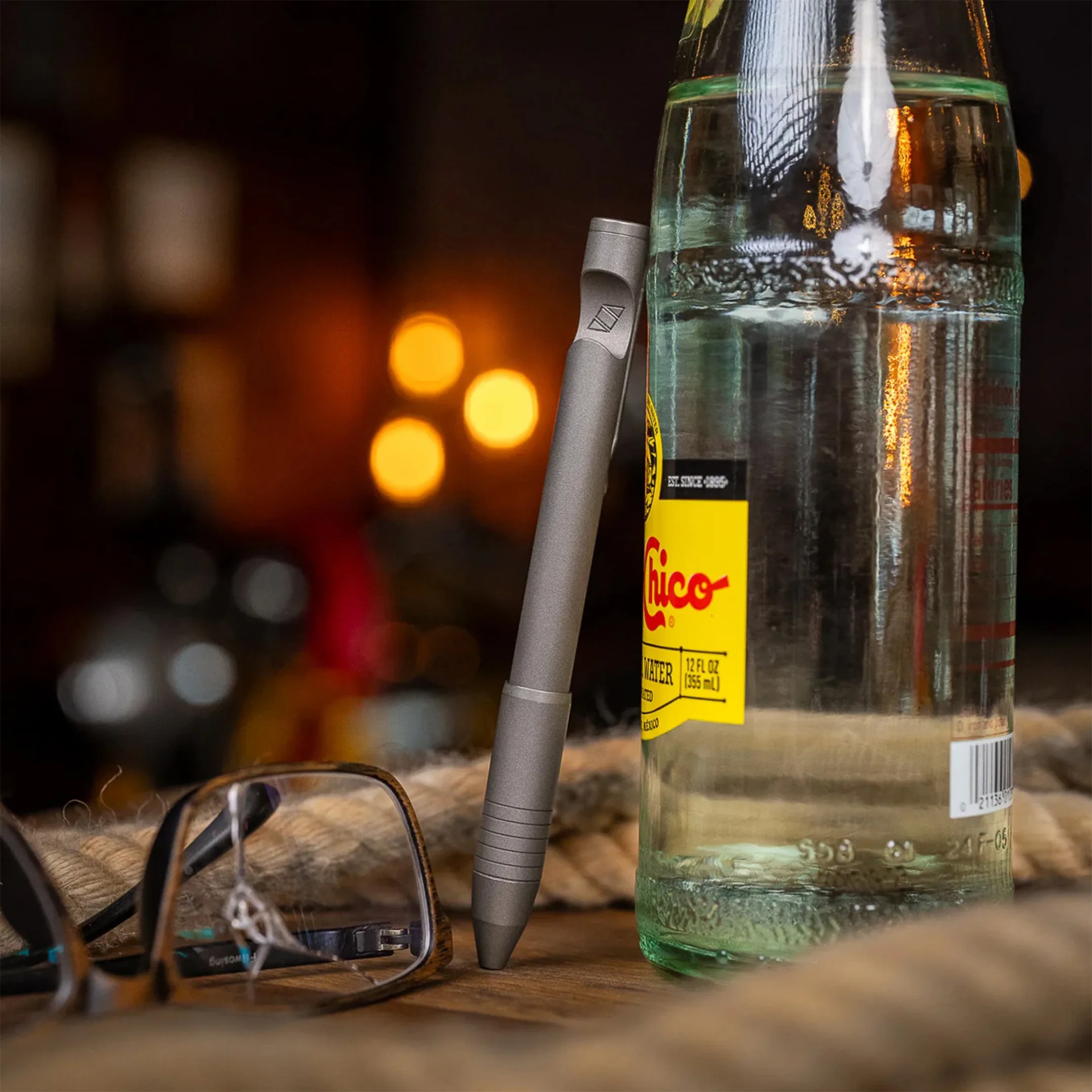 A matte gray pen standing upright, balanced against a glass bottle of Topo Chico sparkling water. The bottle has a yellow label with red and black text. In the foreground, there is a pair of eyeglasses with a tortoiseshell frame resting on a wooden surface. The background is softly blurred with warm, glowing lights.