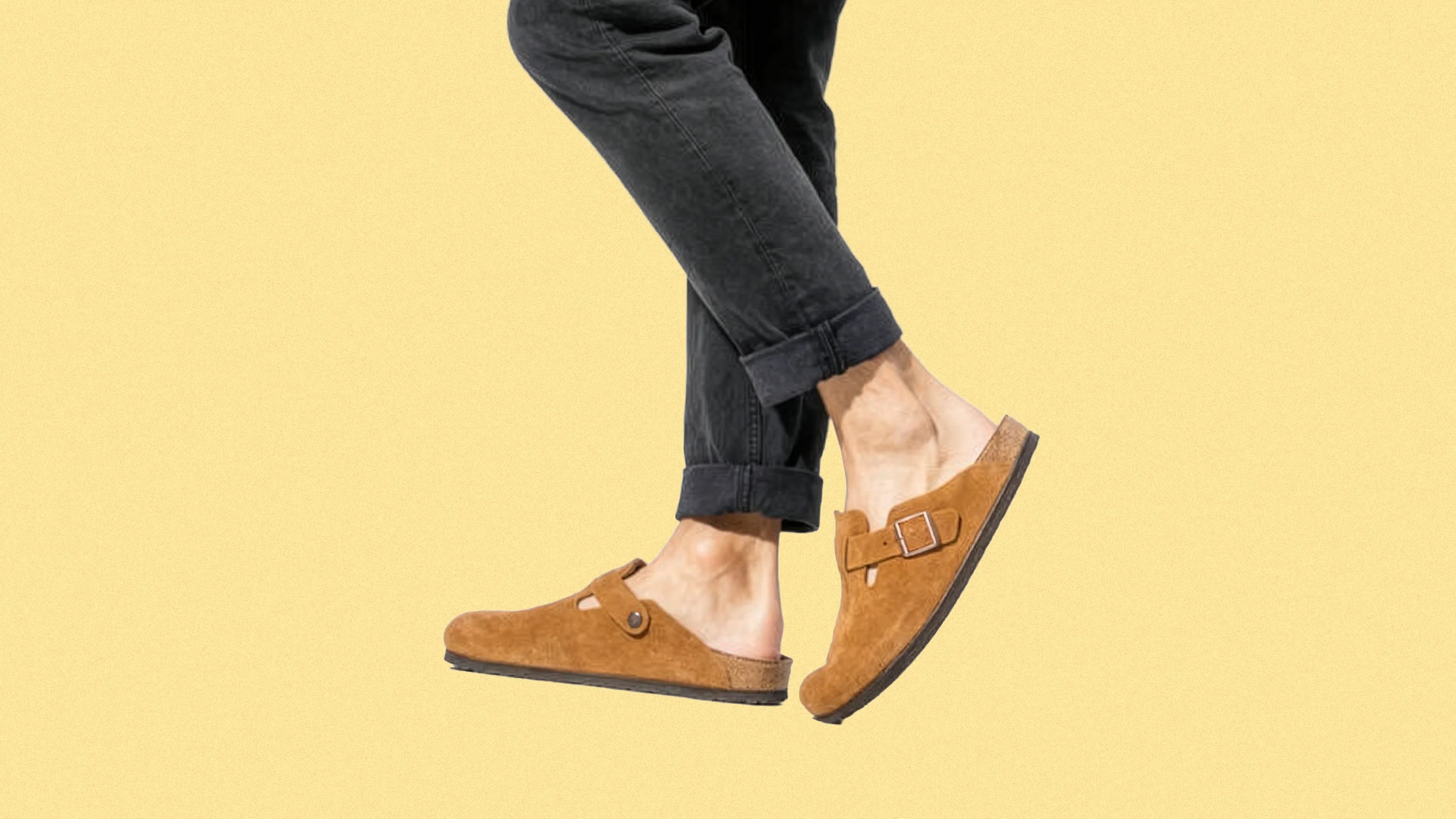 A pair of feet wearing tan suede slip-on clogs with a buckle detail on the side, paired with black rolled-up pants, against a plain light yellow background.