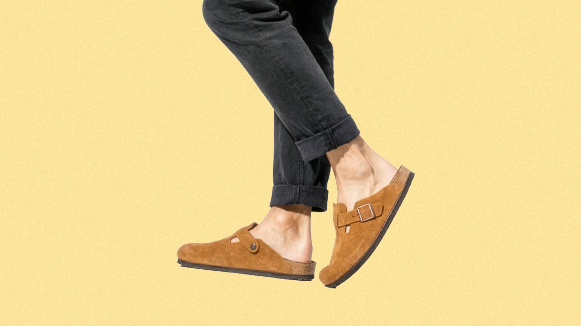 A pair of feet wearing tan suede slip-on clogs with a buckle detail on the side, paired with black rolled-up pants, against a plain light yellow background.
