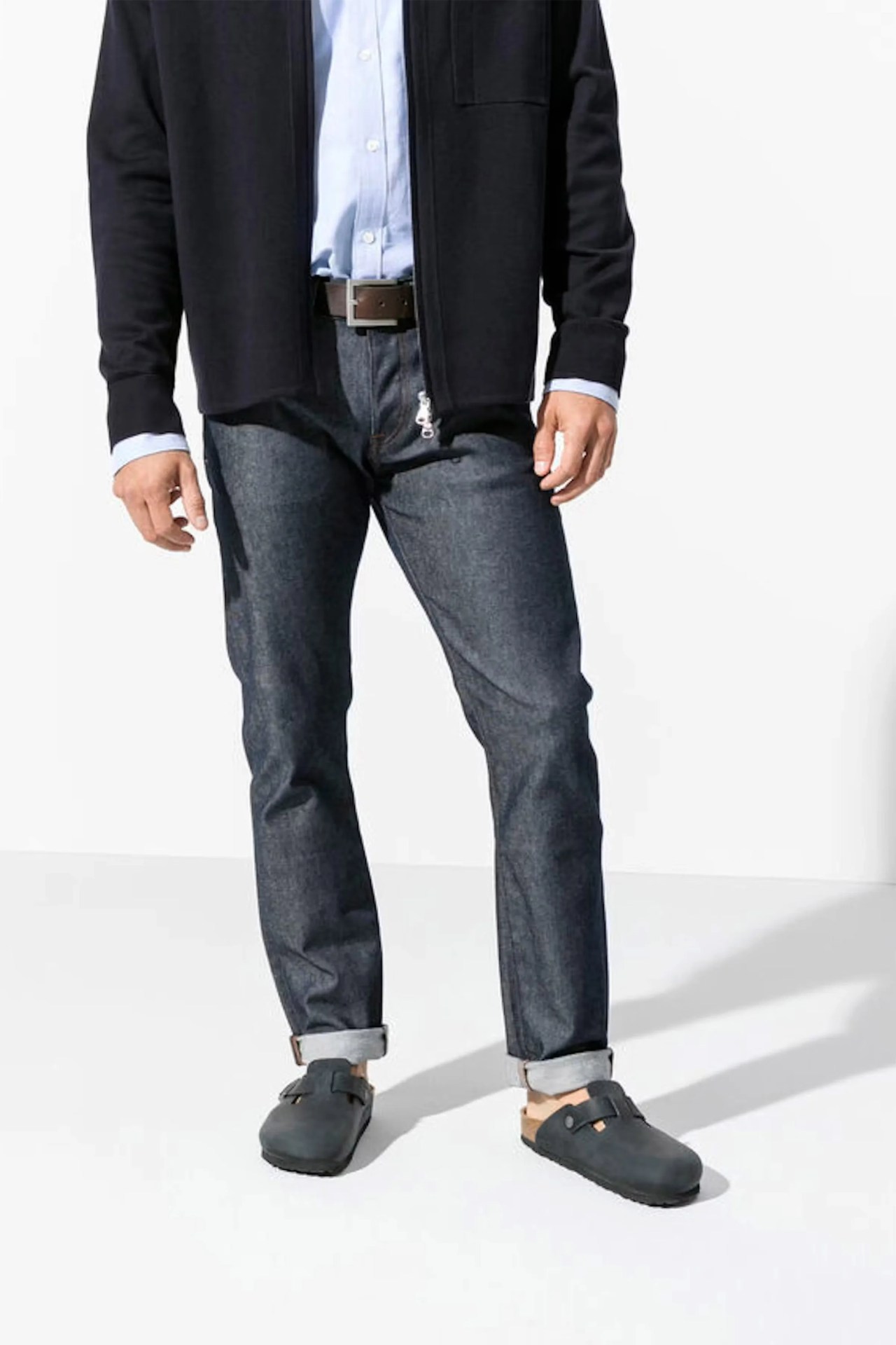 Dark blue denim jeans with a straight fit, rolled up cuffs, paired with a light blue button-up shirt tucked in, a black zip-up jacket, a brown belt, and black slip-on clogs.