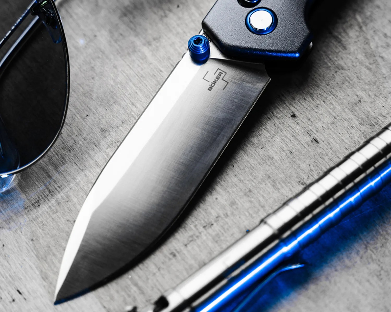 A close-up of a Böker folding knife with a stainless steel blade and a black handle featuring blue accents. The knife is placed on a textured surface next to a pair of dark sunglasses and a metallic pen with a blue reflection.