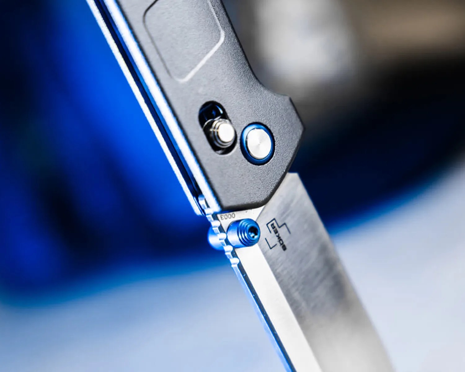 Close-up of a folding knife with a metallic blade and a black handle featuring blue accents, including a blue thumb stud and a blue circular button. The blade has a brushed finish and a logo near the base. The background is blurred with shades of blue and gray.