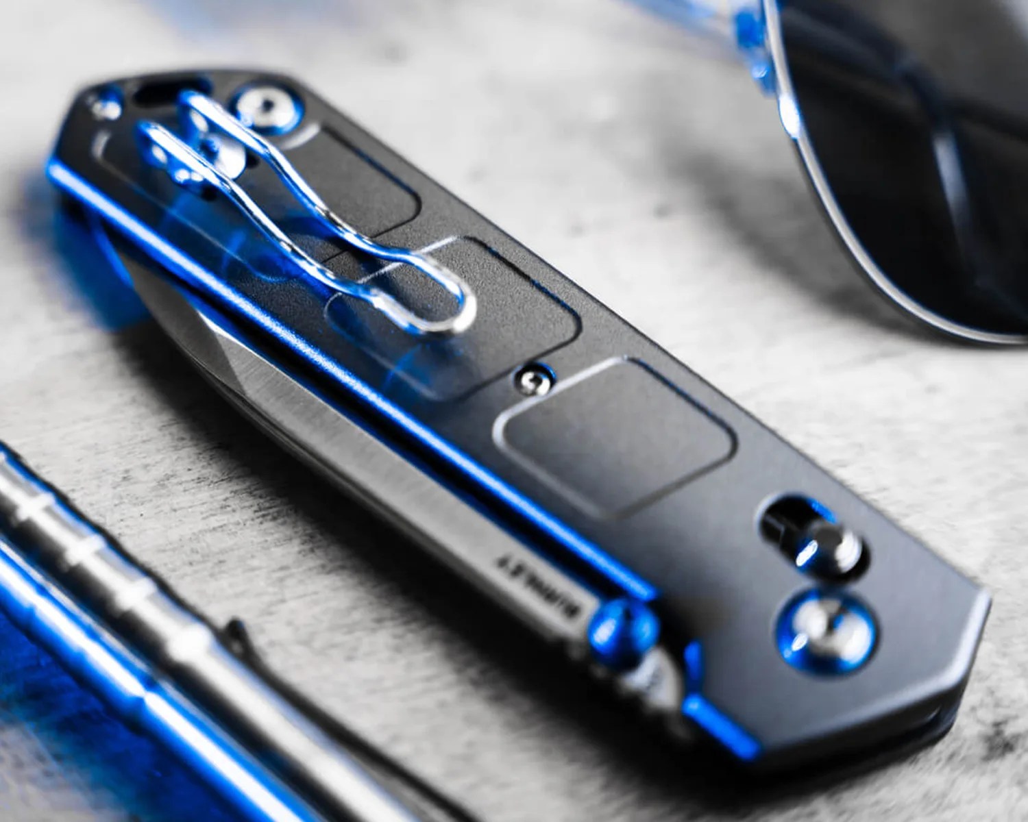 A closed black folding knife with a metal clip attached, resting on a light-colored surface. The knife has a sleek, modern design with visible screws and a partially visible blade edge. Nearby, there is a metallic pen and a pair of dark sunglasses.
