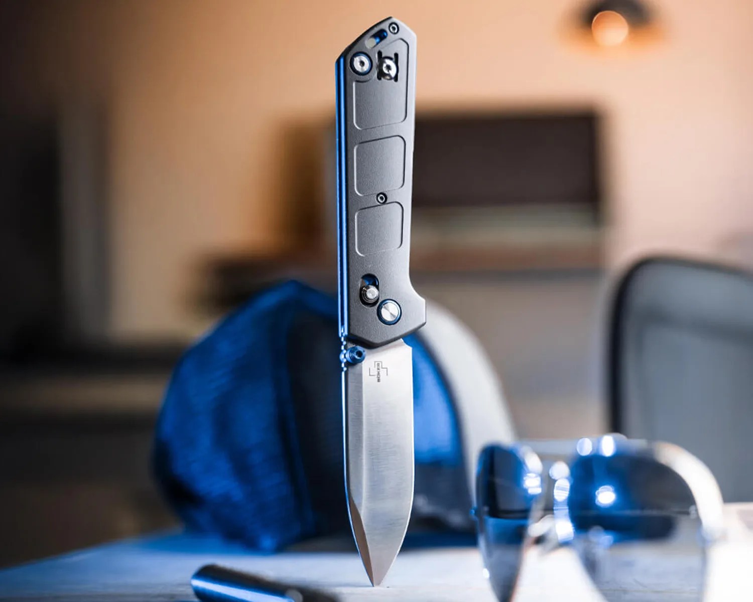 A folding knife with a metallic handle and a sharp, pointed blade standing upright on a surface. The blade has a logo near the base, and the background is blurred with a blue cloth and some reflective objects nearby.