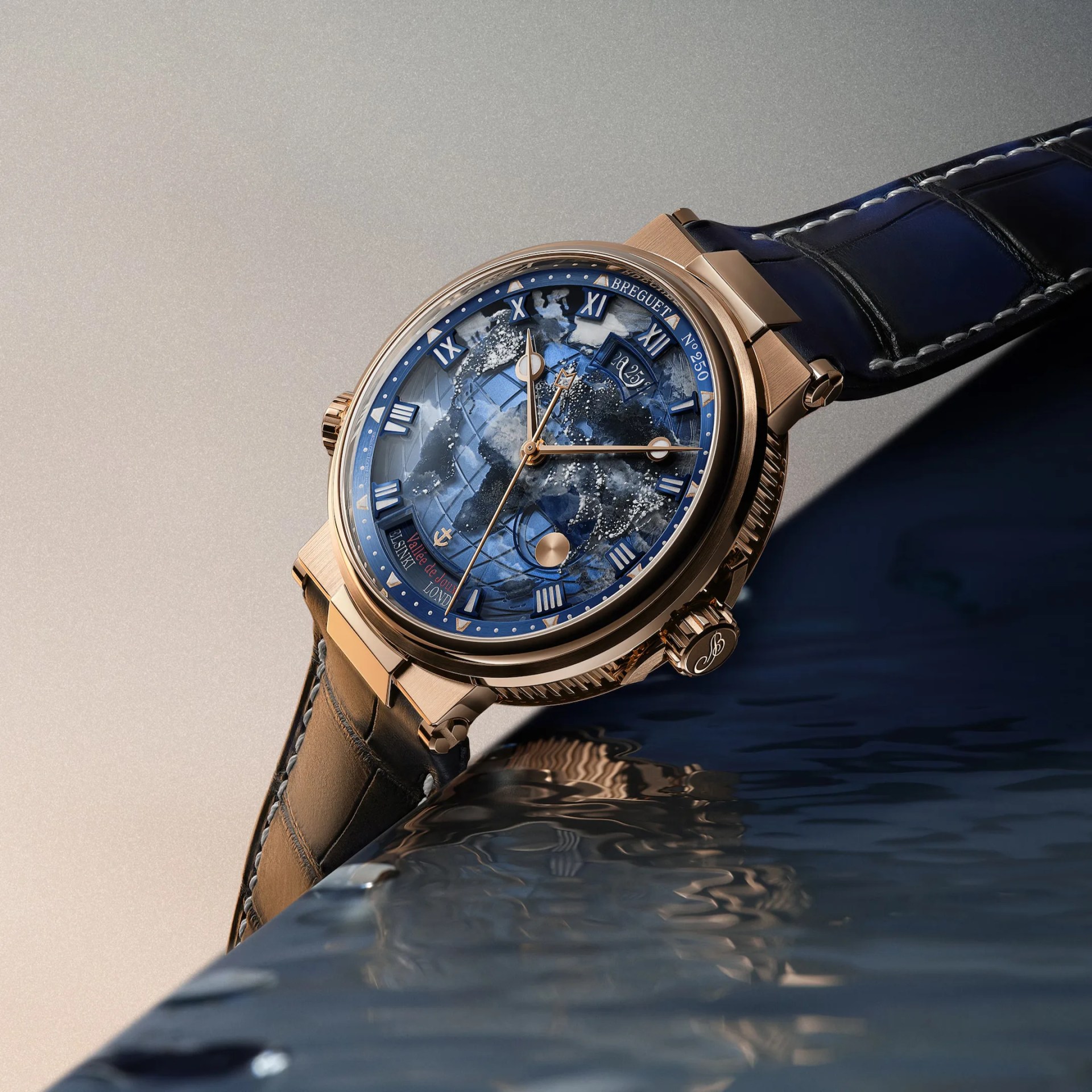 Luxury wristwatch with a gold case and a deep blue dial featuring a detailed world map design. The watch has Roman numeral hour markers, gold hands, and a date window at the 12 o'clock position. It is paired with a dark blue leather strap with white stitching. The crown is engraved with a decorative "B." The watch is positioned on a reflective surface with a gradient background.